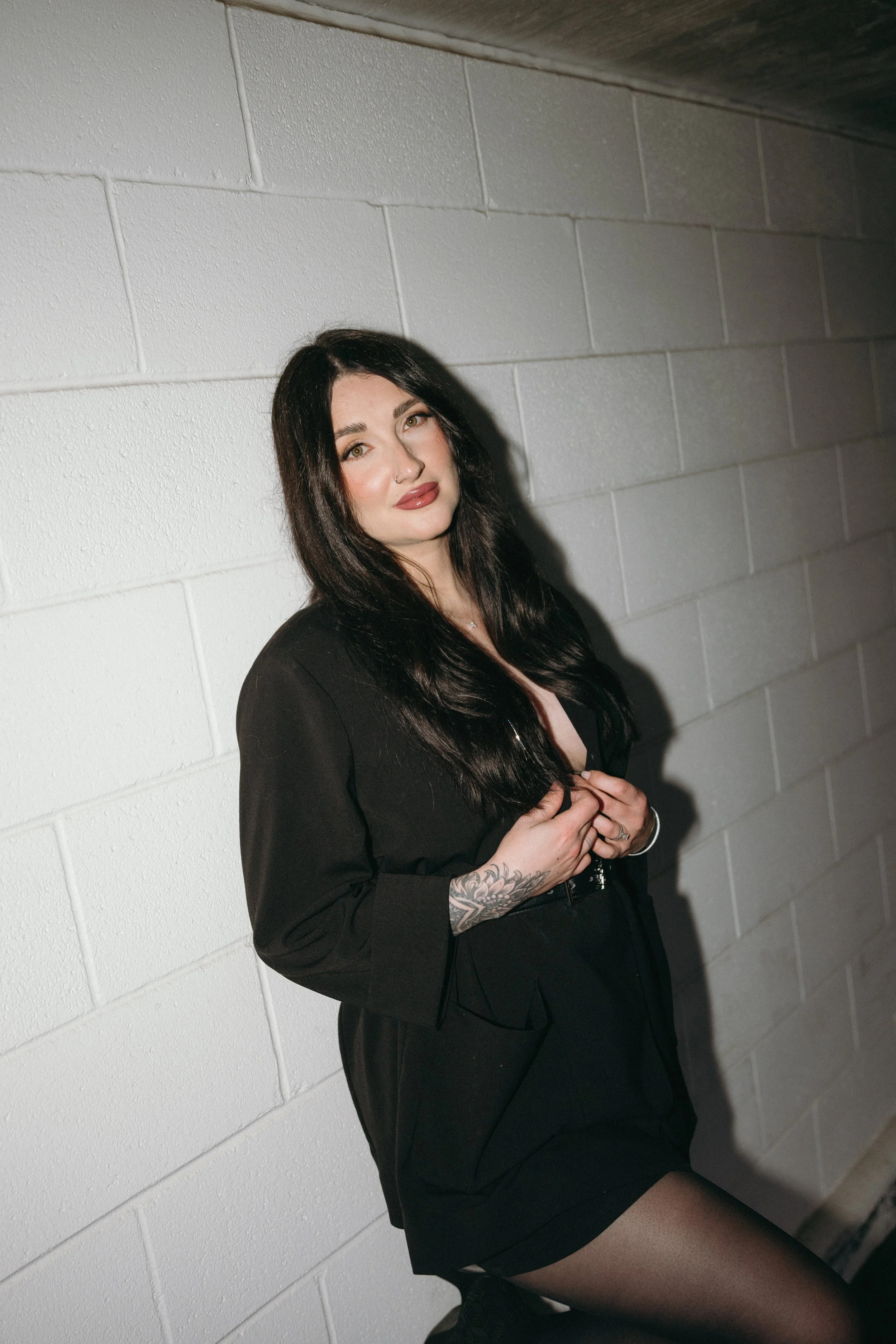A woman with long dark hair, wearing a black blazer and skirt, posing against a white cinder block wall.