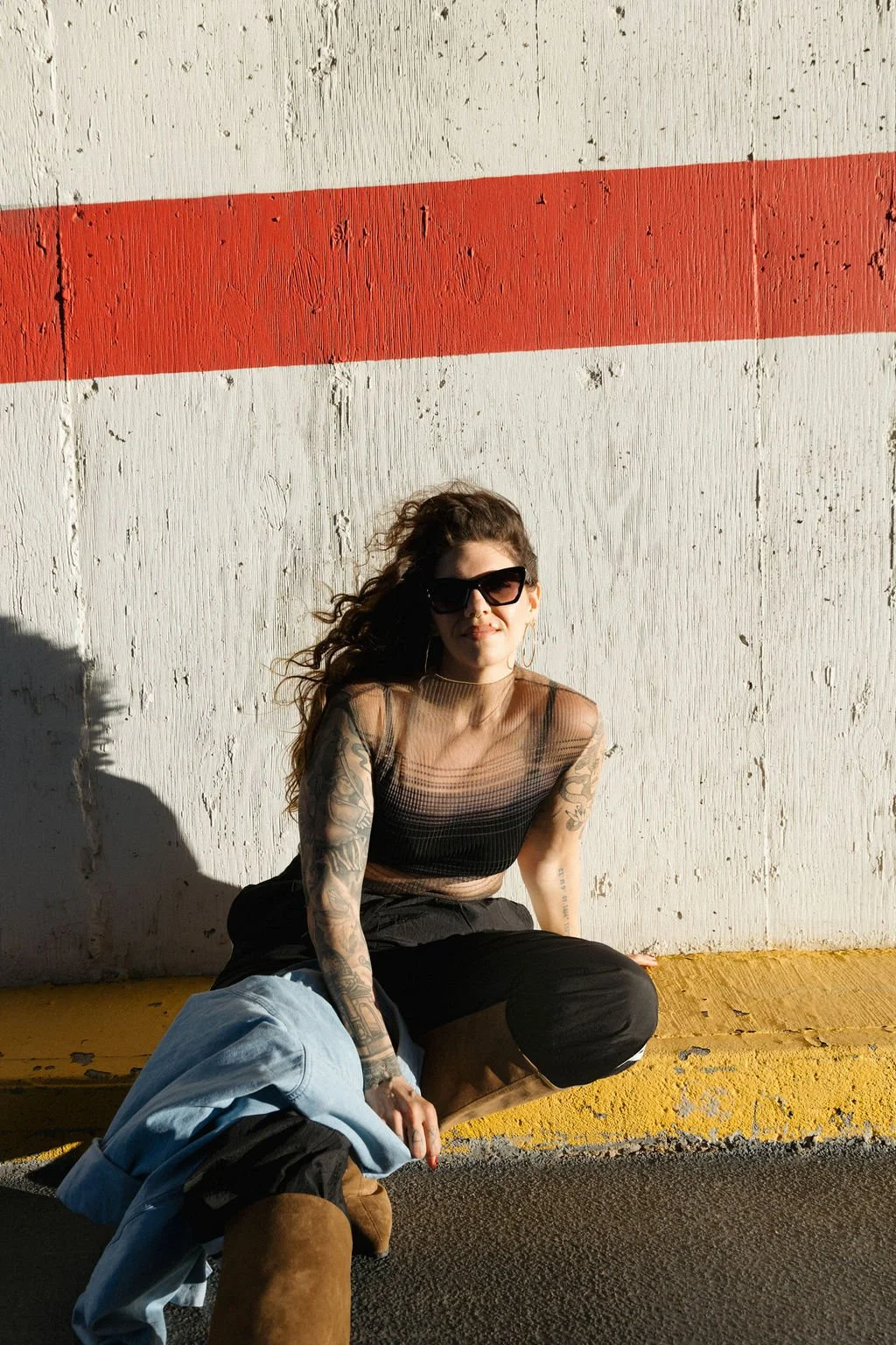 A young woman with curly brown hair leaning against a brick wall in an outdoor urban setting, holding a string of beads in one hand and a small bottle in the other, wearing a black blazer and black leather pants.