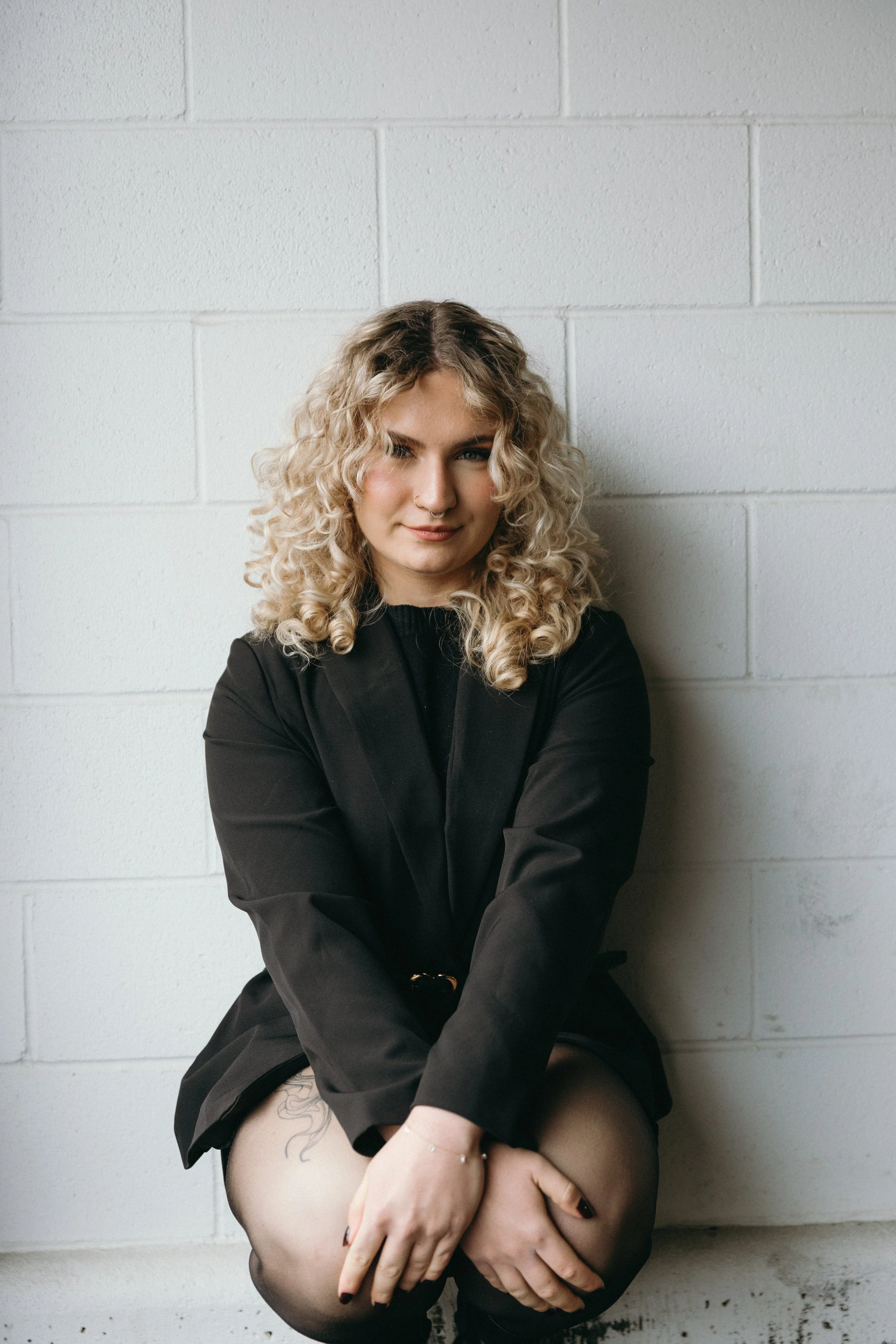 A woman with curly blonde hair sitting against a white brick wall, wearing a black blazer and sheer black tights.