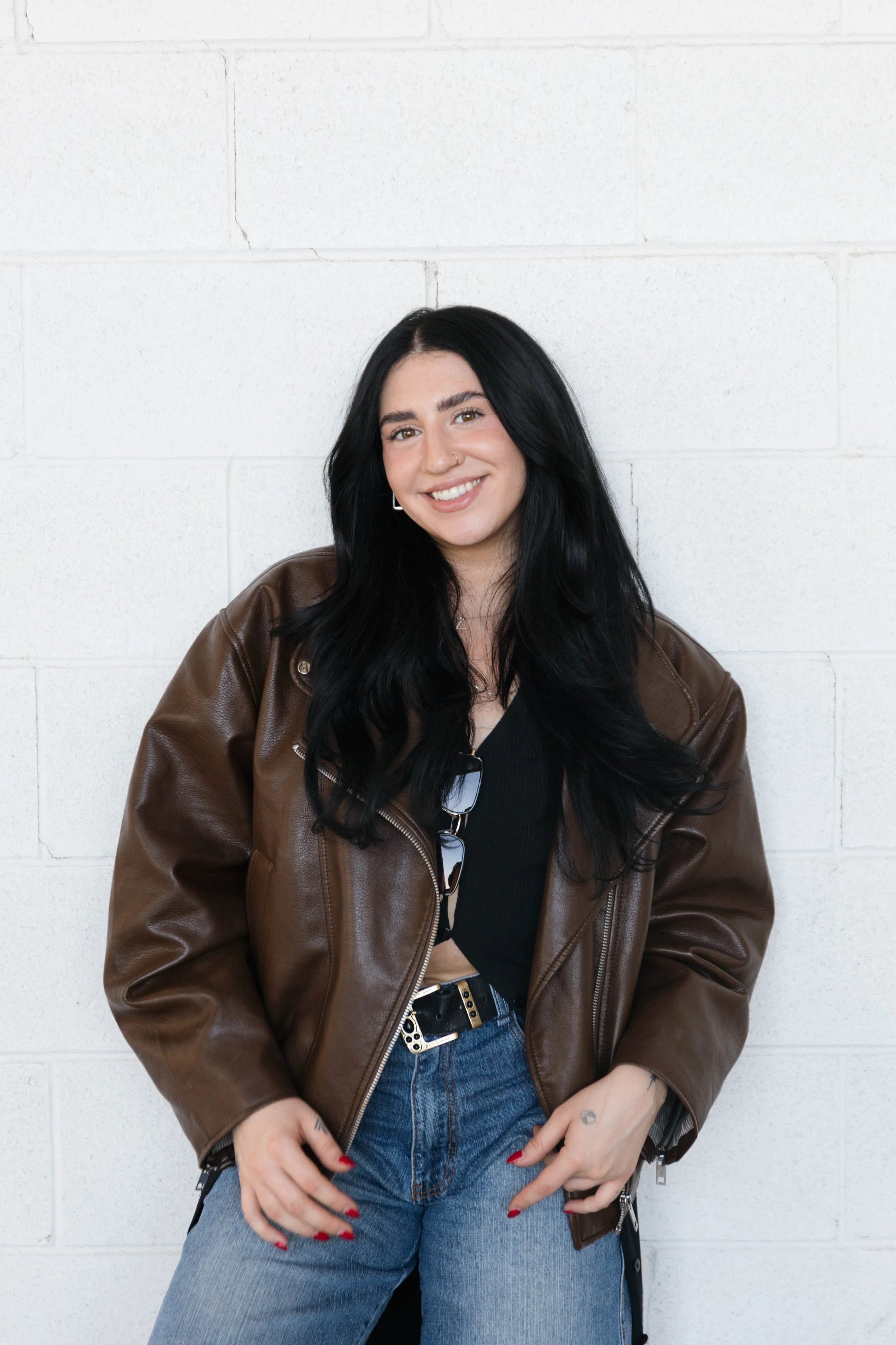 A smiling woman with long, dark, wavy hair is sitting on a railing under an overpass, wearing a black blazer and pants.