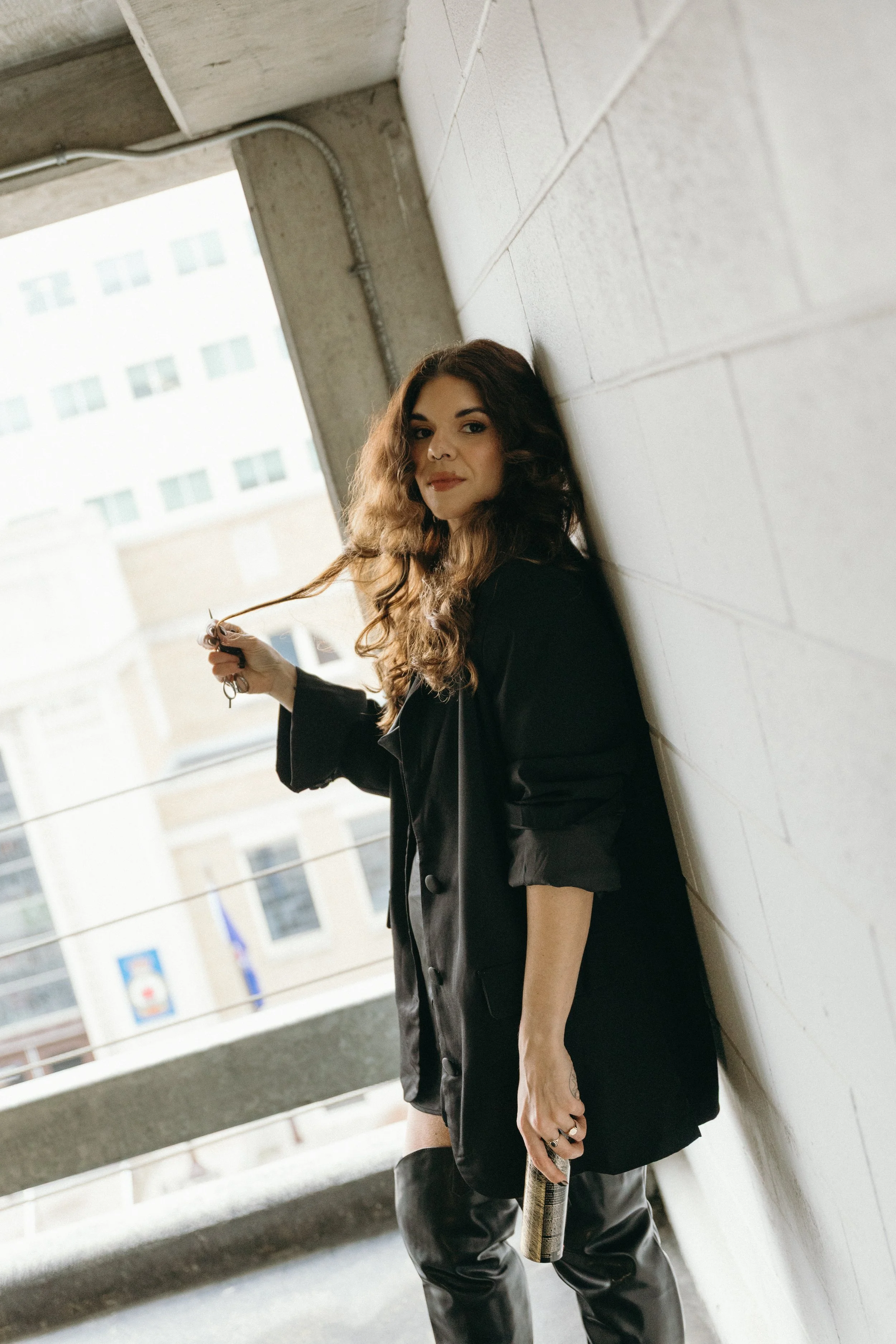 A young woman with curly brown hair leaning against a brick wall in an outdoor urban setting, holding a string of beads in one hand and a small bottle in the other, wearing a black blazer and black leather pants.