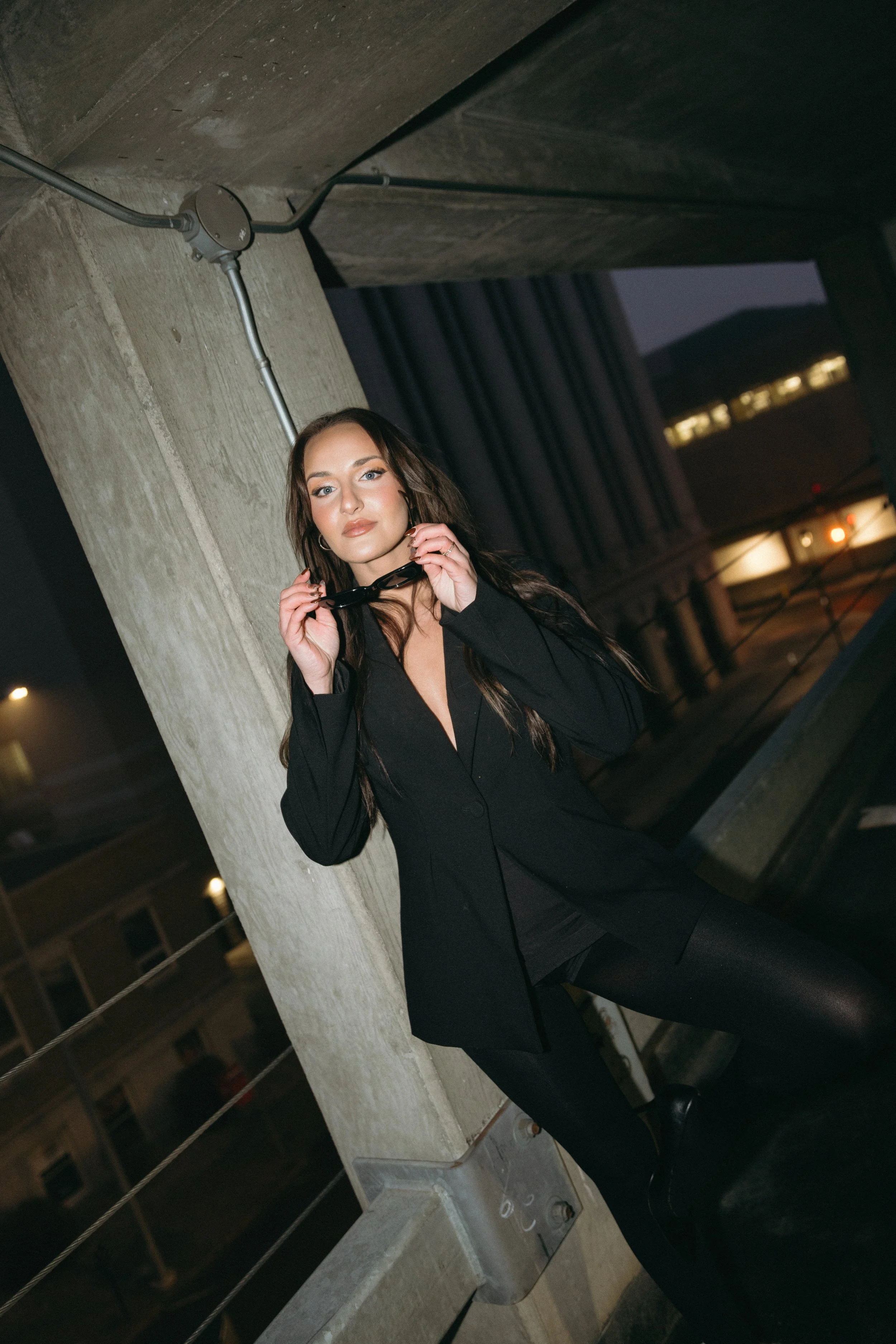 A woman with long dark hair holding black sunglasses, wearing a black blazer and leggings, standing on a balcony at night with urban buildings and lights in the background.