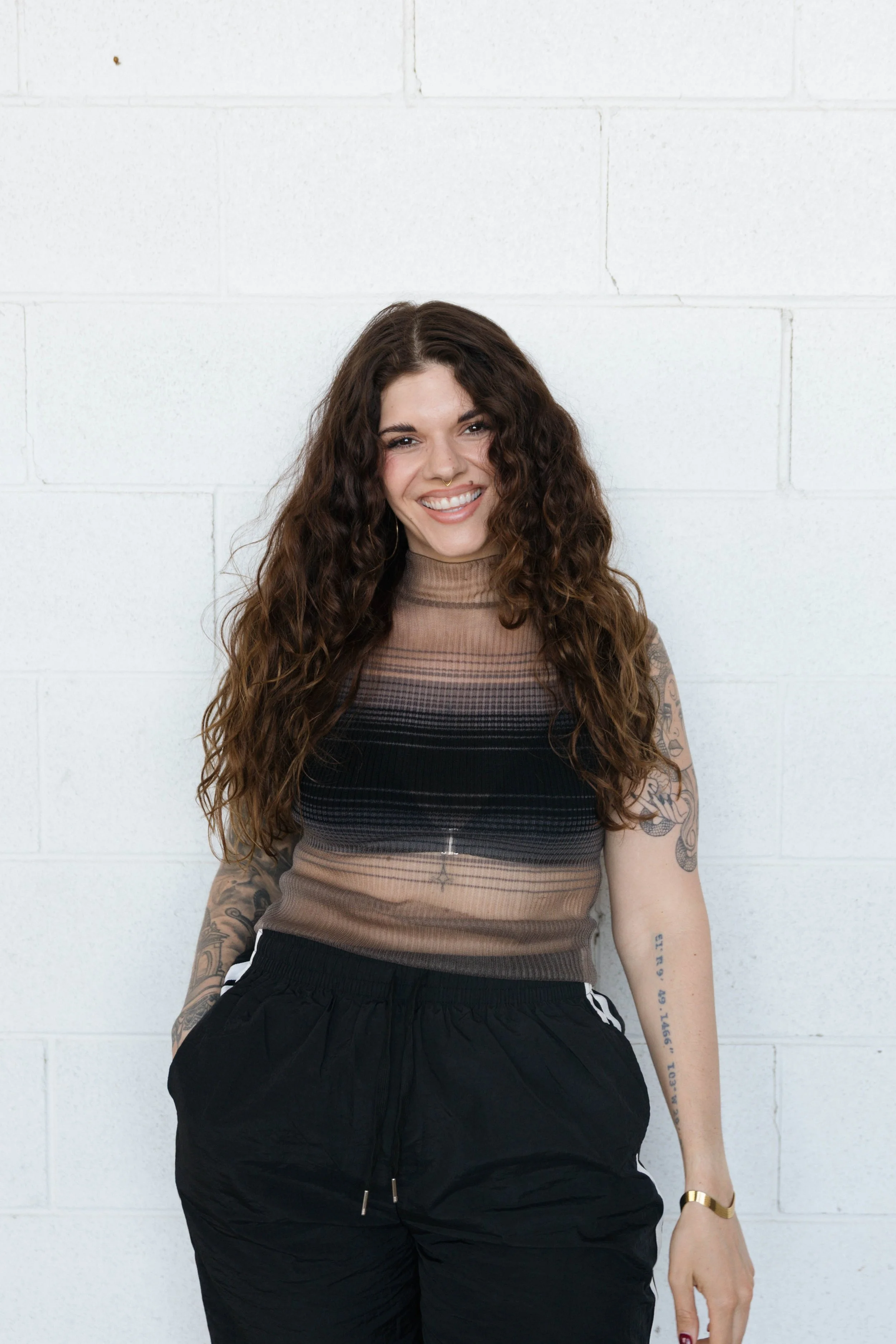 A young woman with curly brown hair leaning against a brick wall in an outdoor urban setting, holding a string of beads in one hand and a small bottle in the other, wearing a black blazer and black leather pants.