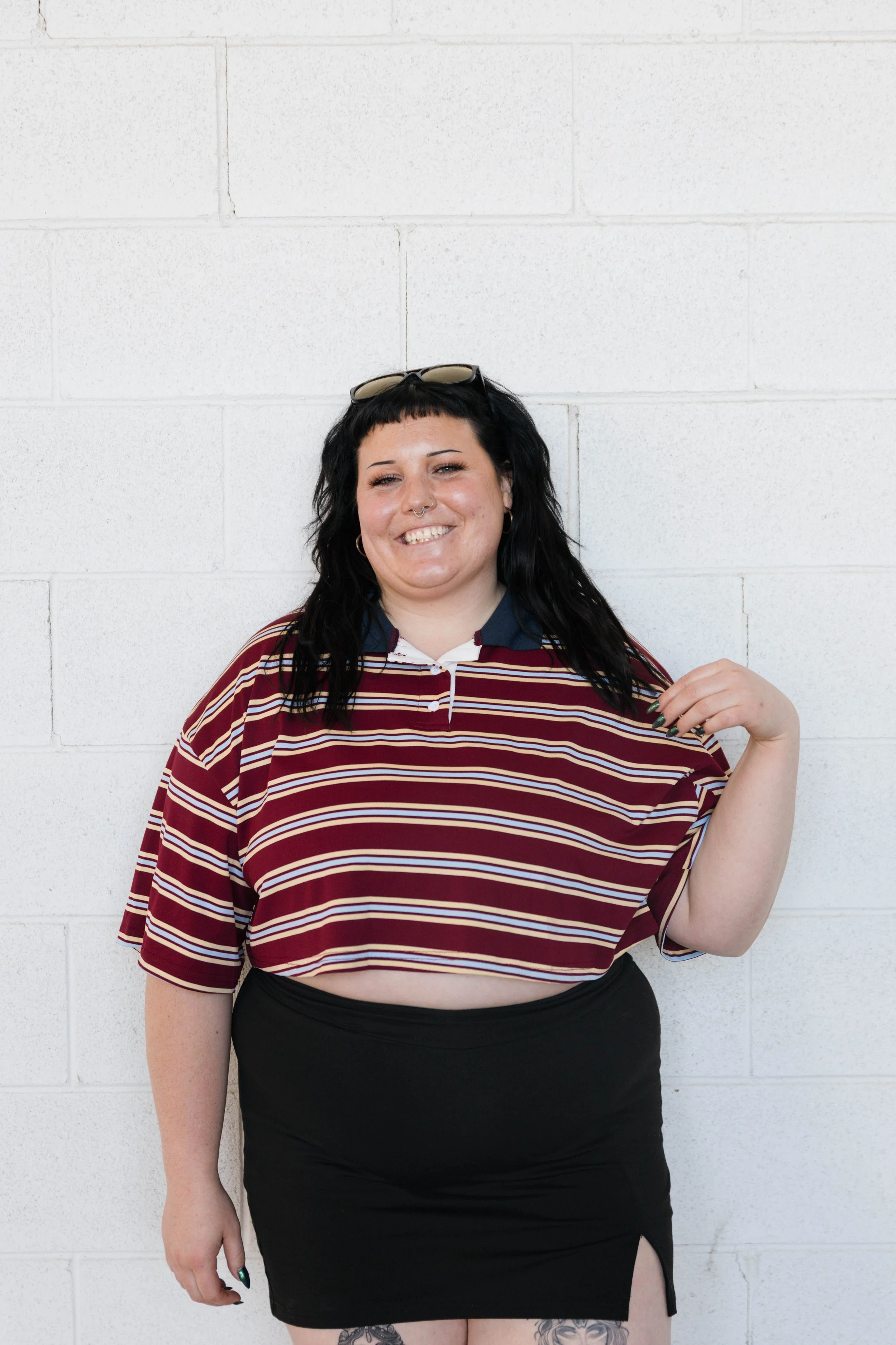 A woman with long dark hair and a septum piercing, smiling and wearing a black ribbed sweater, standing against a white brick wall.