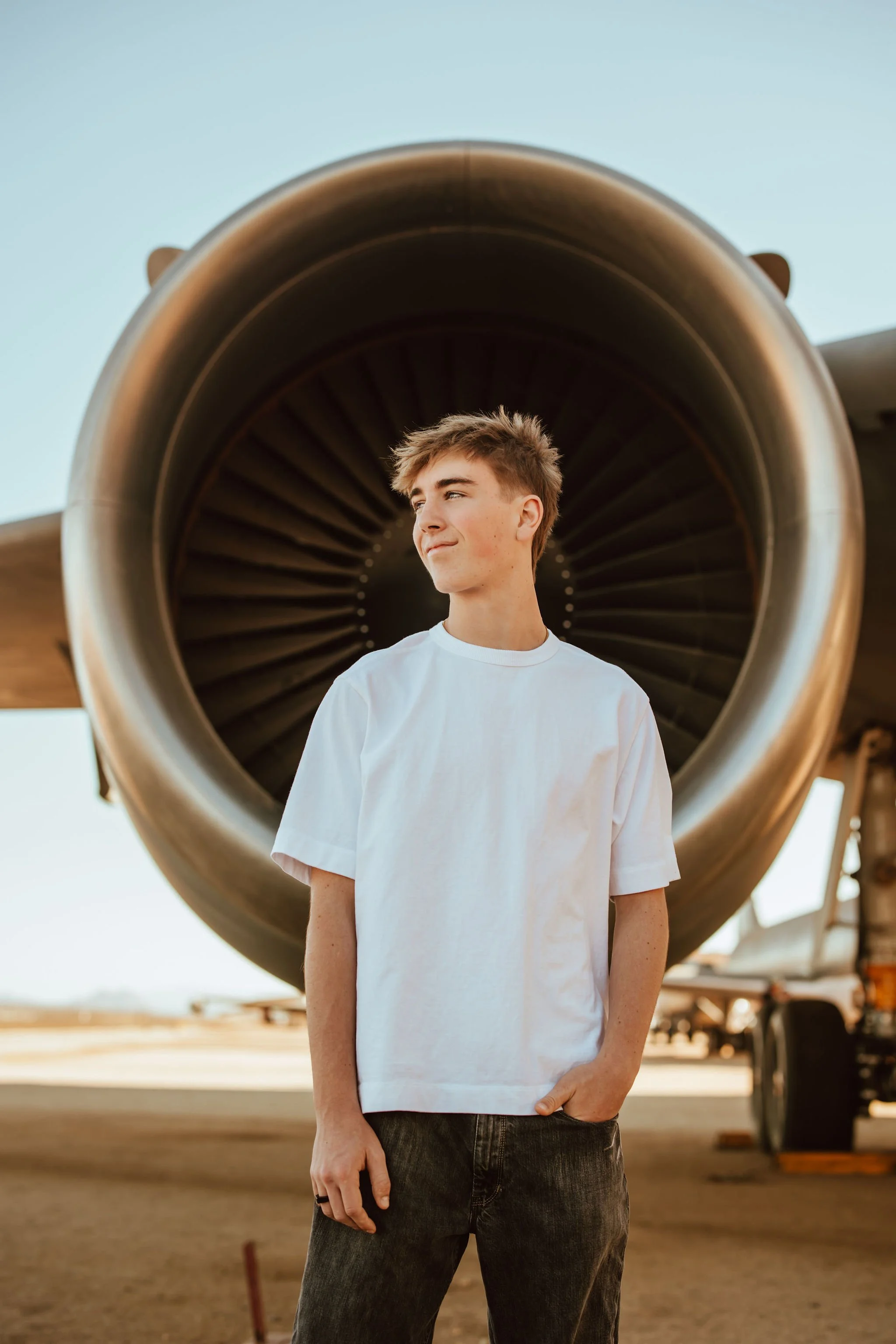 A young man in a white T-shirt and black jeans standing in front of an airplane engine at an airport or airfield during sunset.