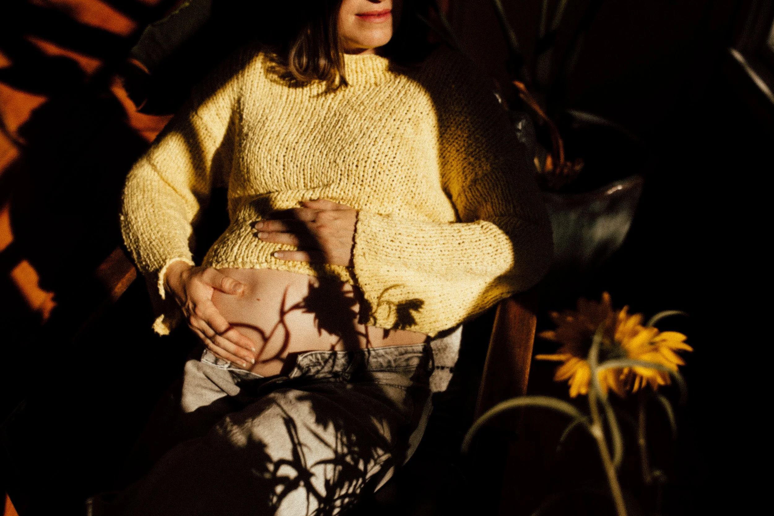 A pregnant woman lying on a wooden bench, wearing a yellow sweater and having her hands on her belly in a dimly lit space with shadows of flowers.
