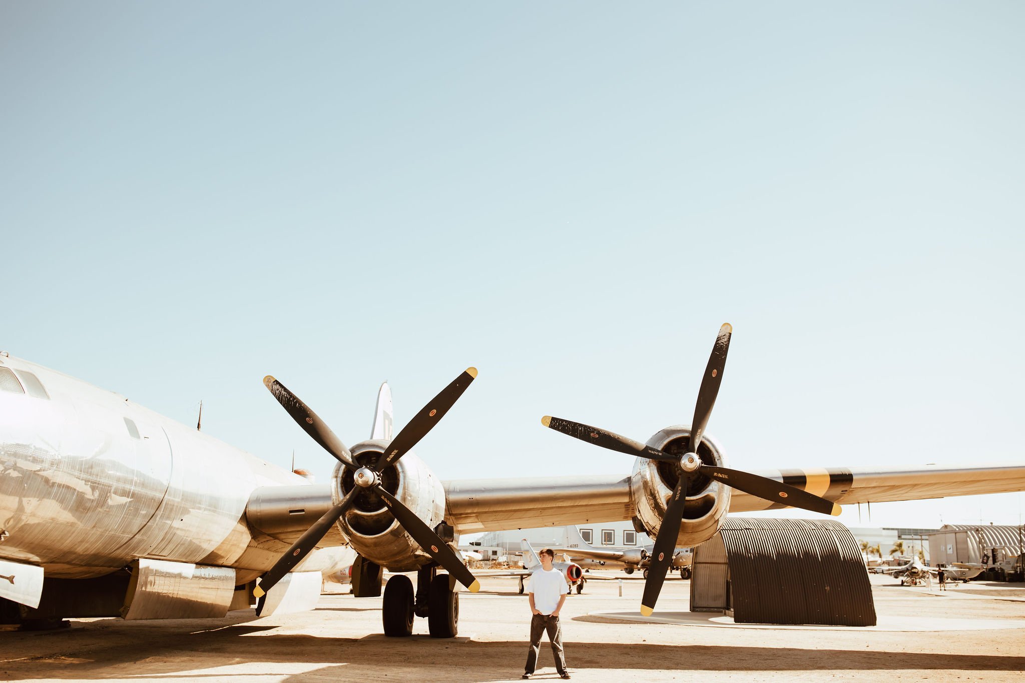 A person standing under a vintage twin-engine propeller airplane at an outdoor aircraft museum, with other airplanes and hangars in the background.