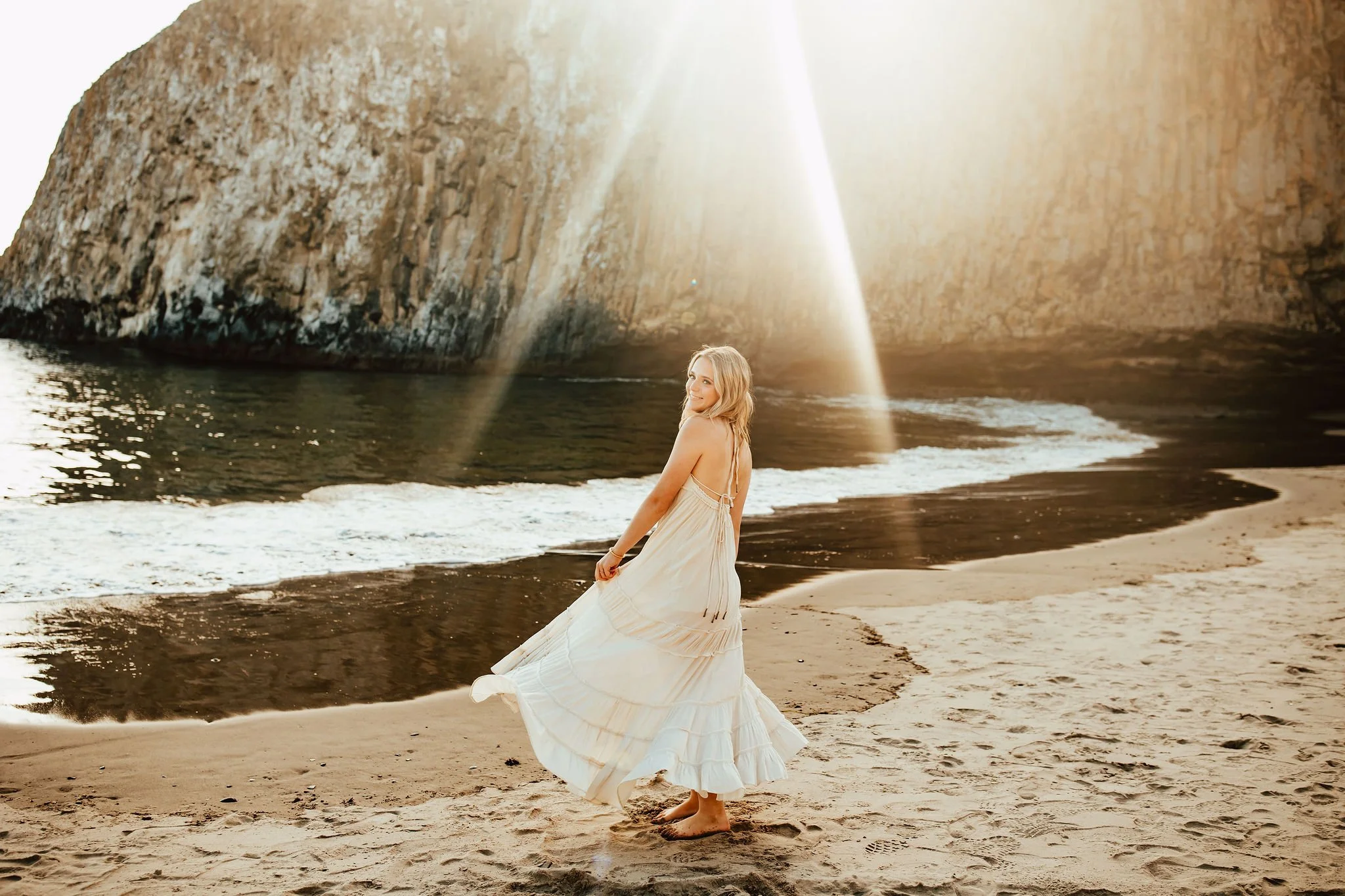 A woman in a white flowing dress standing on a sandy beach near the water with cliffs in the background, bathed in warm sunlight.