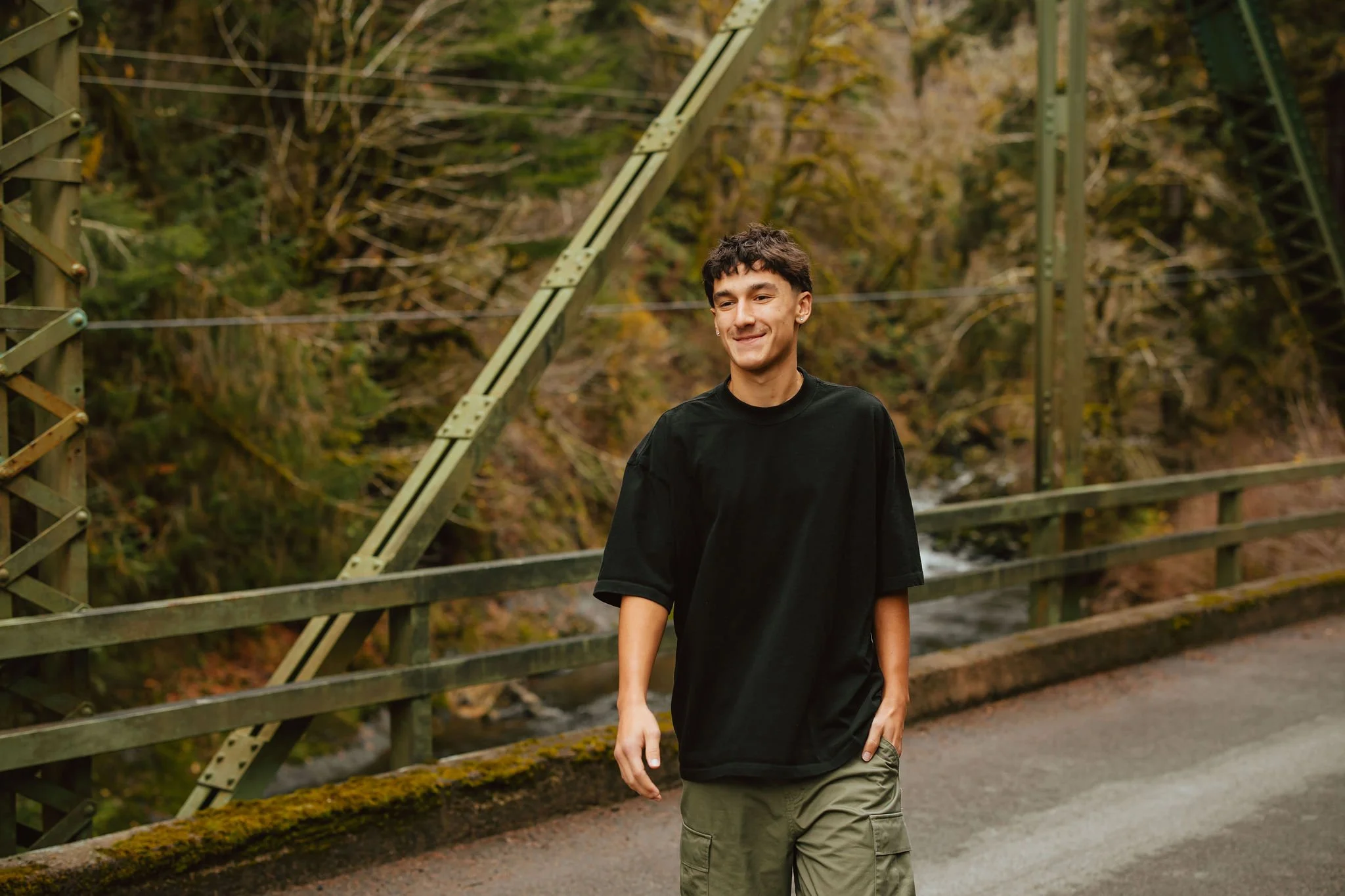 A young man wearing a black t-shirt and olive cargo shorts walking across a bridge in a wooded area.