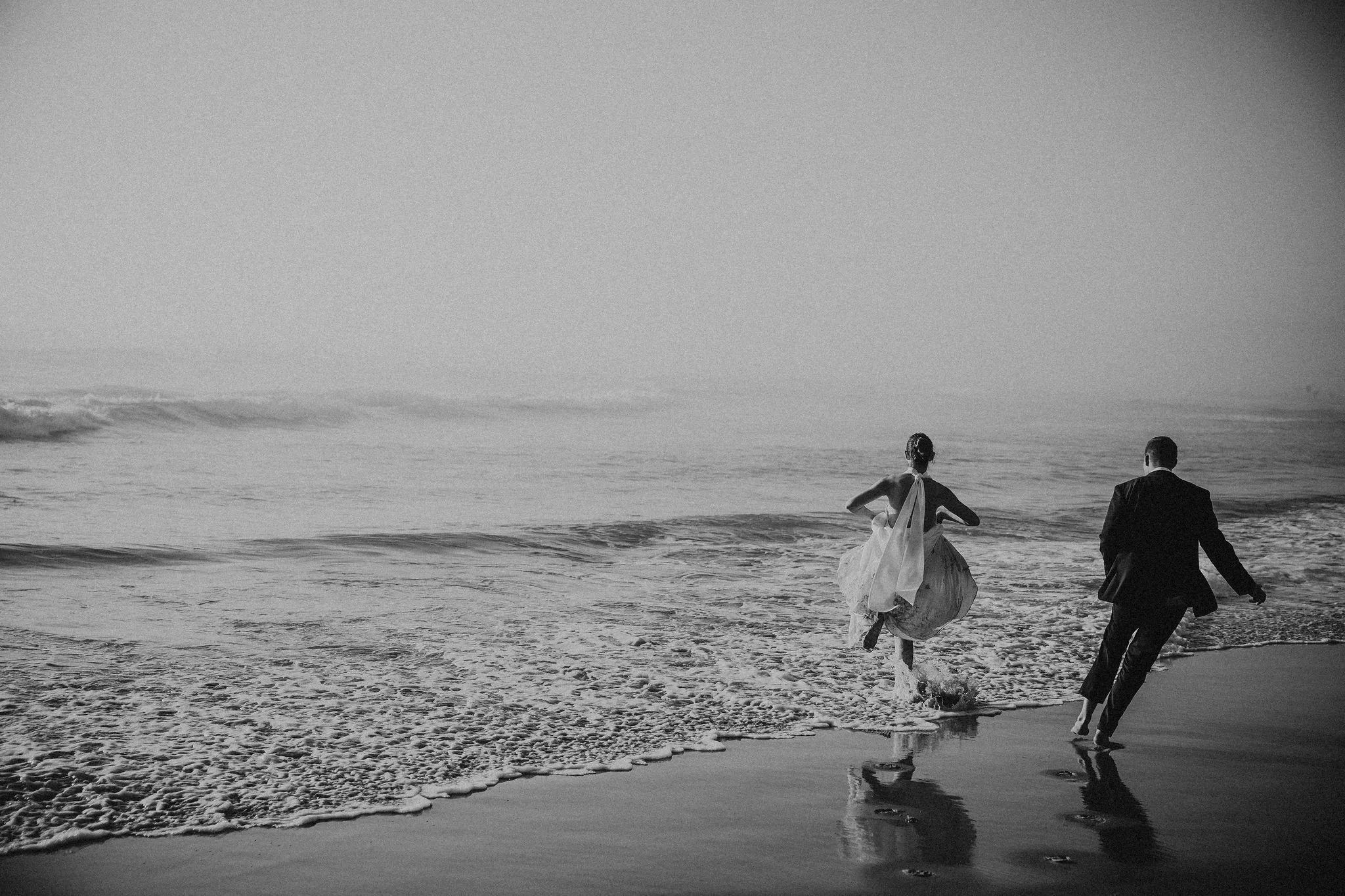 A black and white photo of a bride and groom running along the shoreline at the beach, with waves touching their feet.