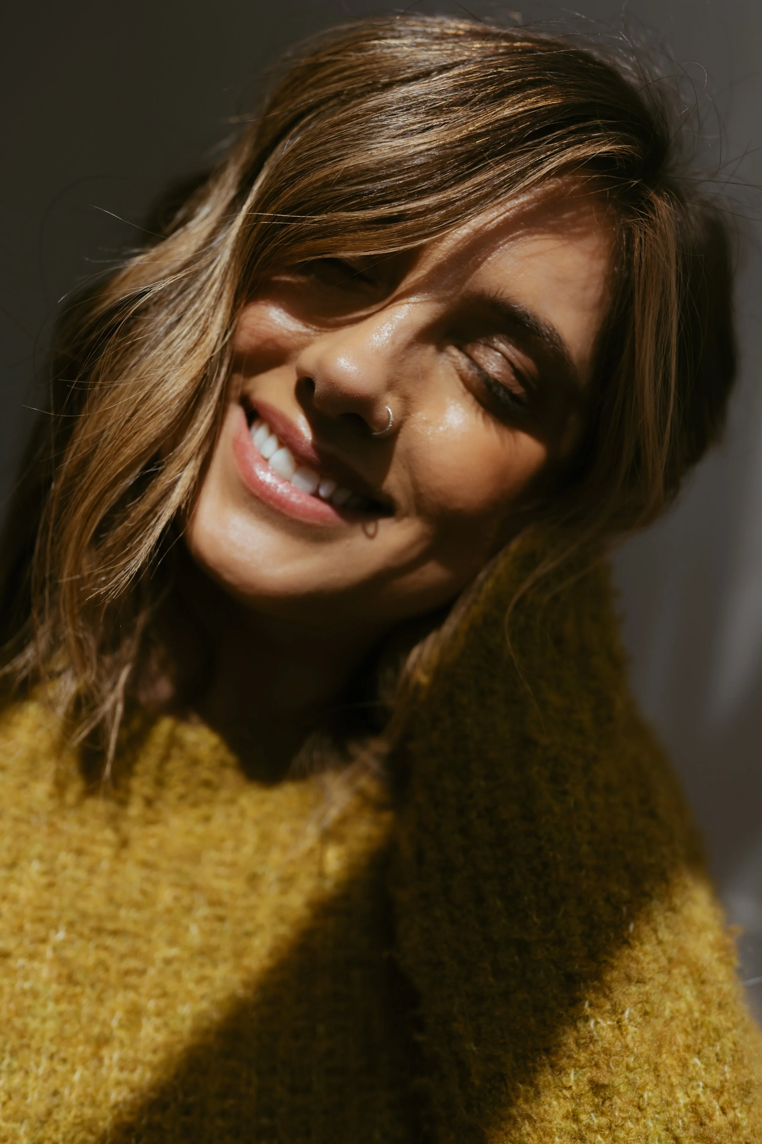 A young woman with light brown hair, smiling with her eyes closed, wearing a mustard-colored sweater, in natural light.