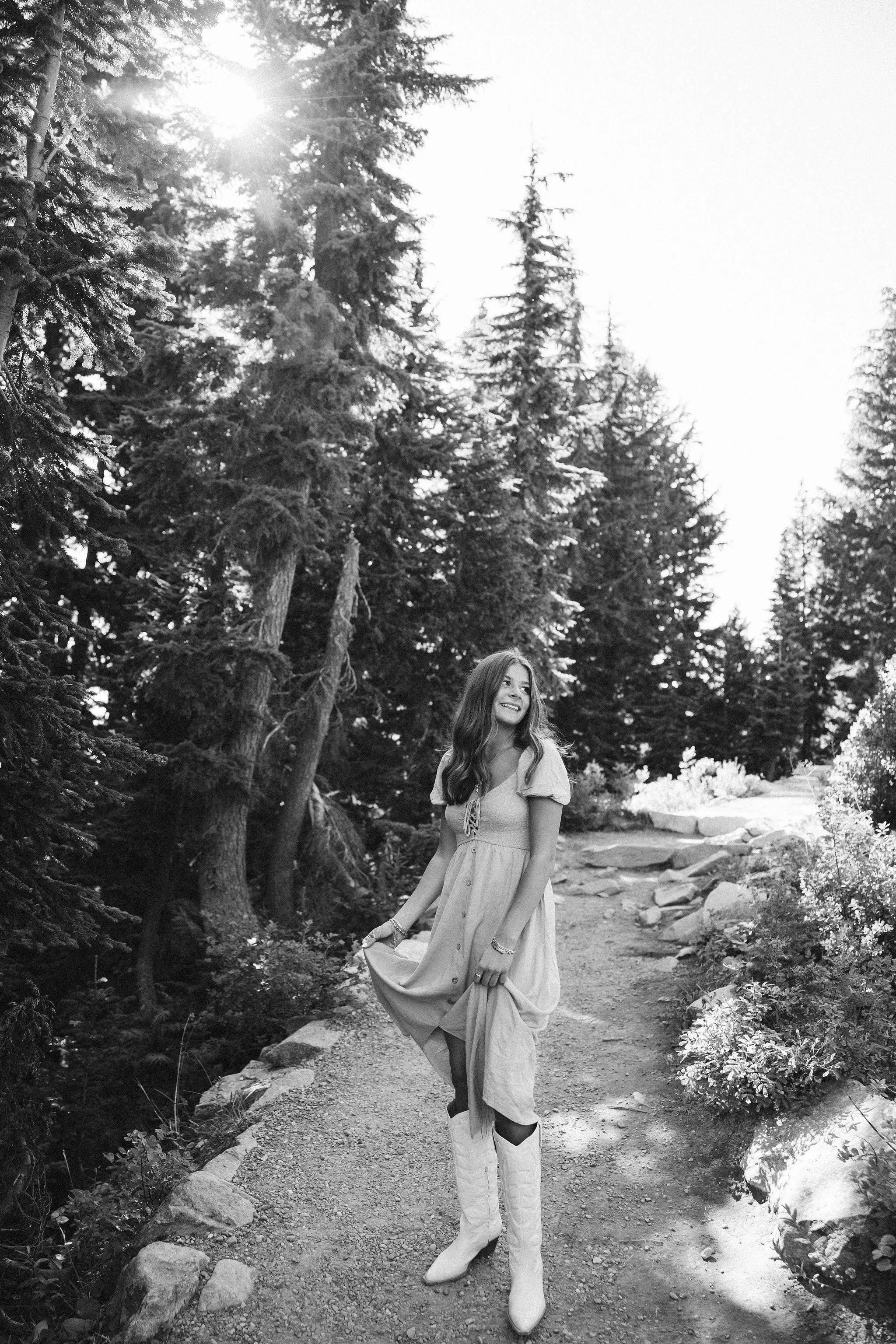 A woman in a light-colored dress and white cowboy boots walking on a dirt trail surrounded by tall pine trees, with sunlight shining through the branches, in a forest setting.