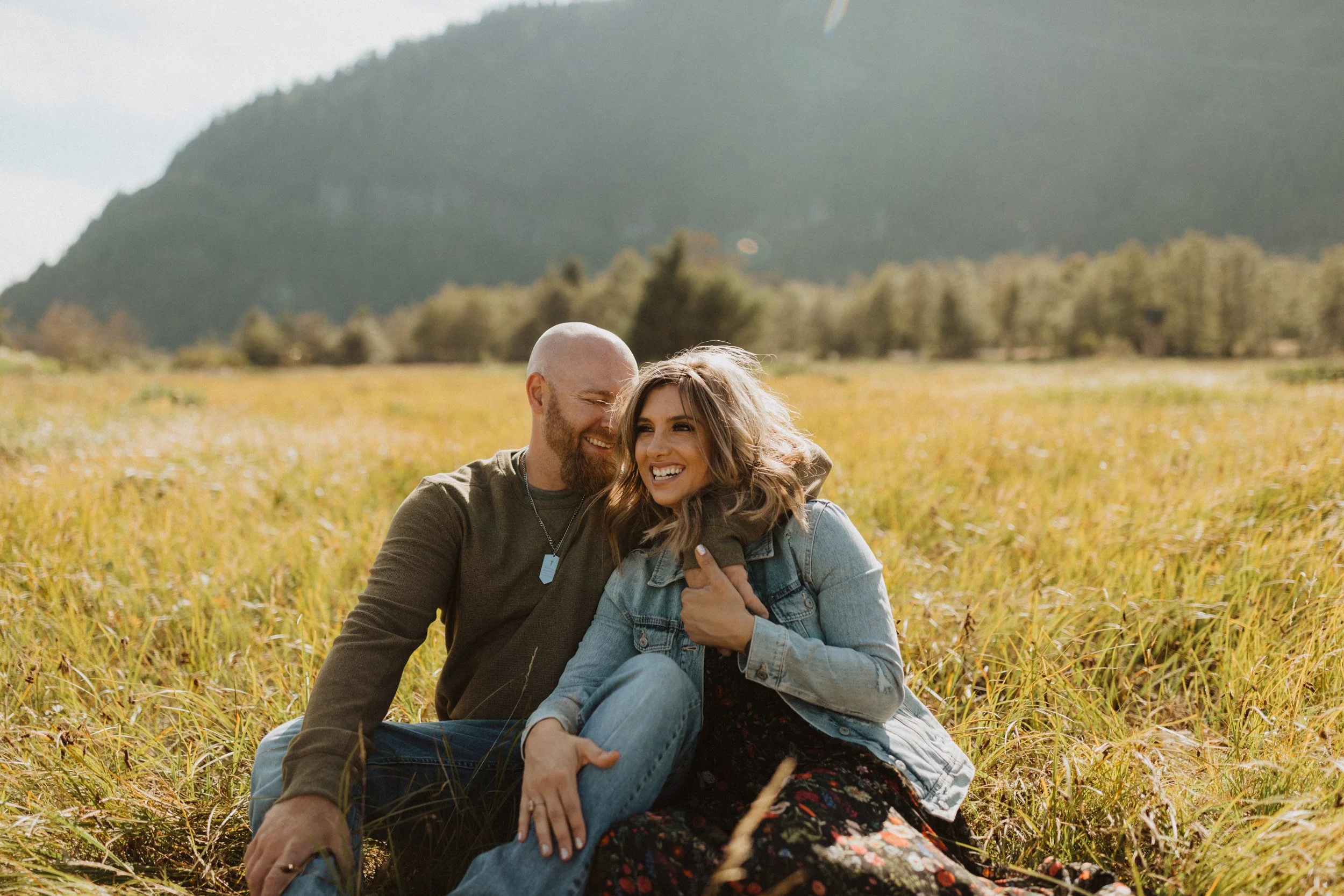 A happy couple sitting on the grass in a field, smiling and laughing, with mountains and trees in the background.