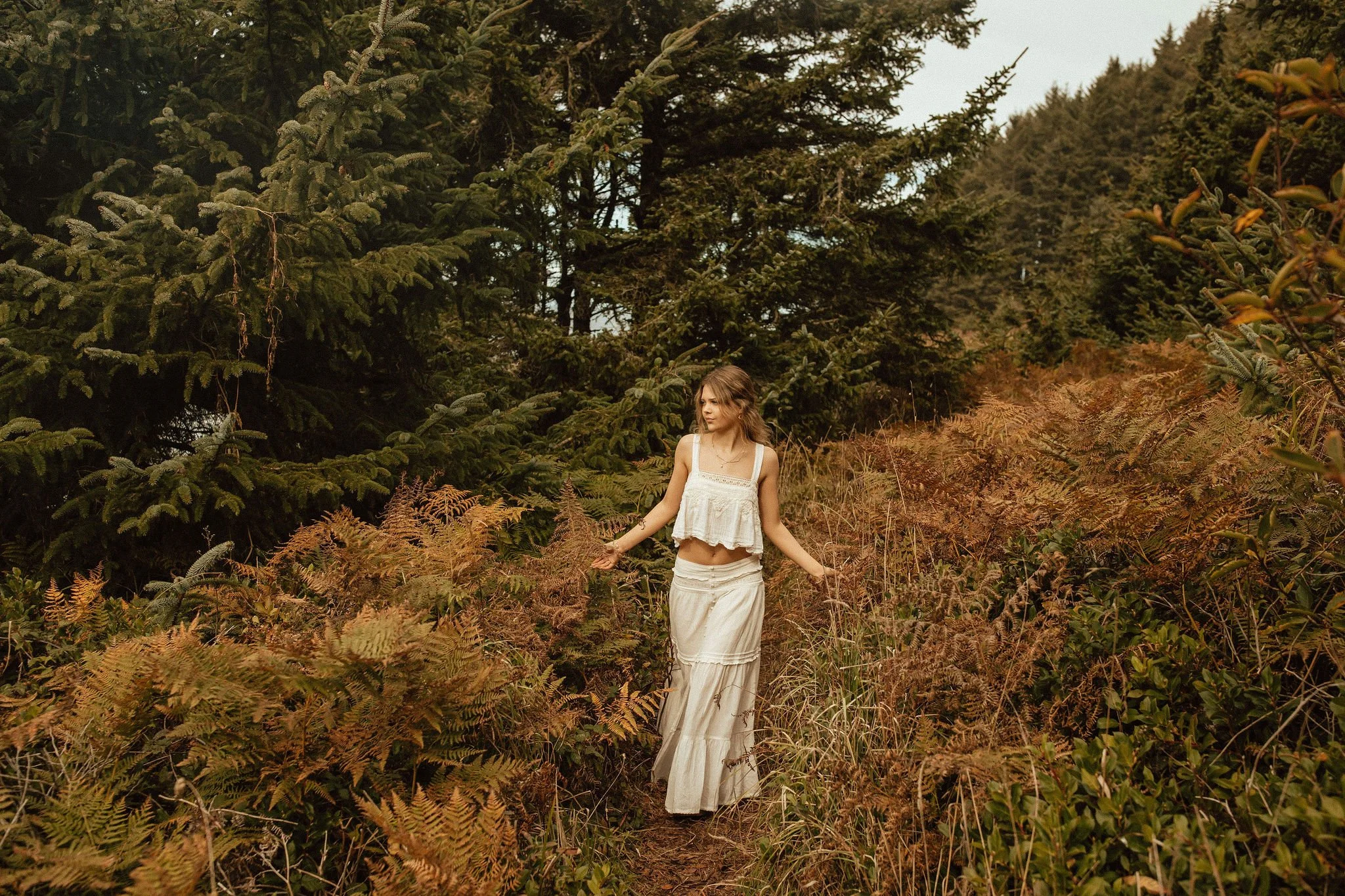 A young woman in a white crop top and long skirt walking along a narrow dirt trail in a forested area with lush green and brown ferns and tall pine trees.