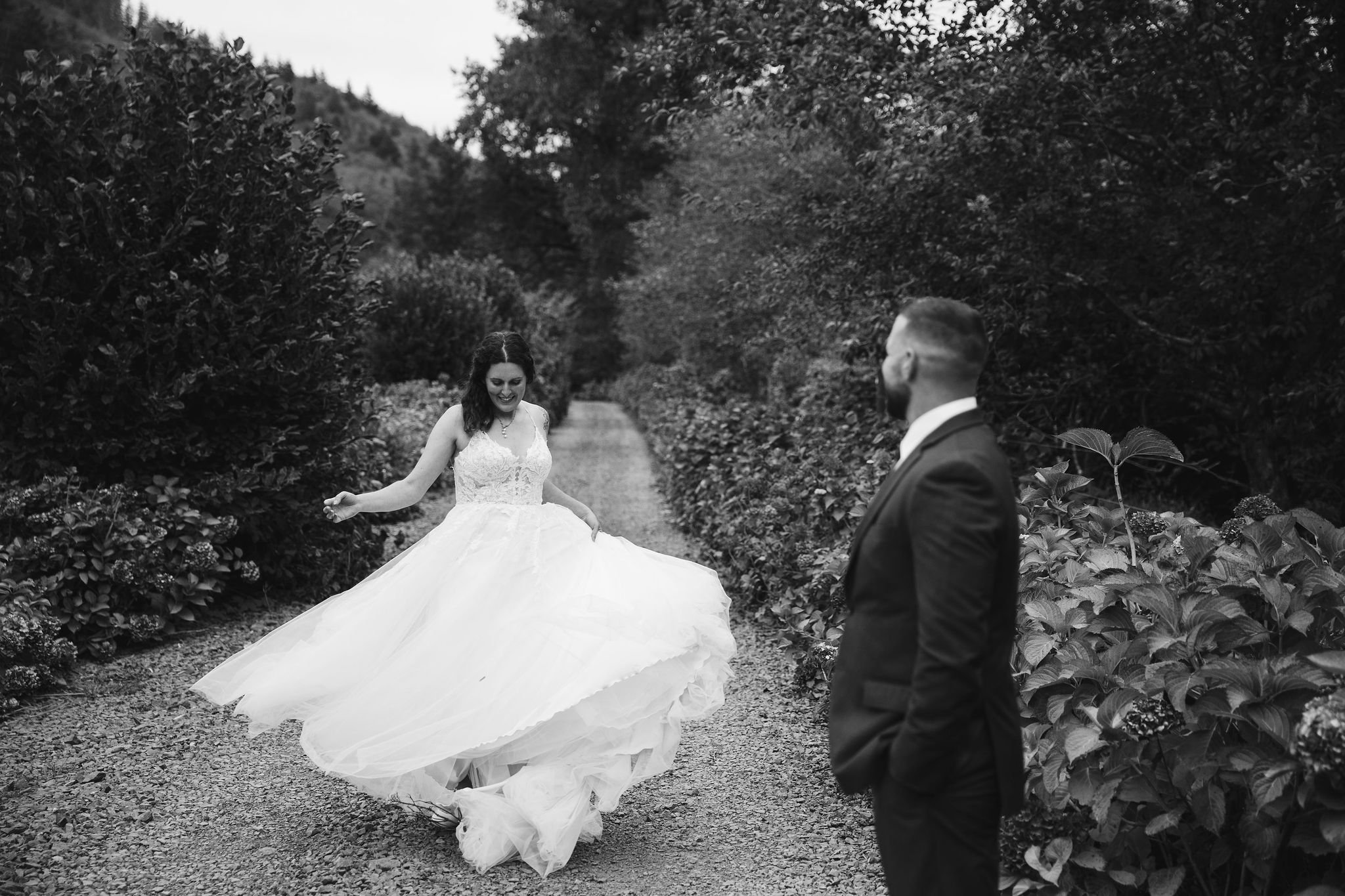 A black and white photo of a bride in a wedding dress dancing on a gravel path, while a groom watches her in a suit. The scene is outdoors with trees and bushes lining the path.