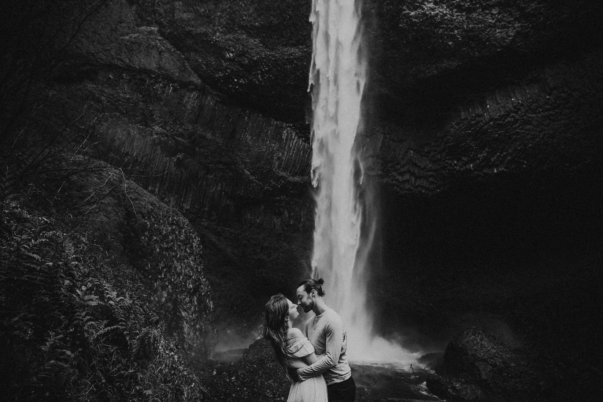 A couple in love stands close together in front of a waterfall, embracing and looking into each other's eyes in a black-and-white photograph.