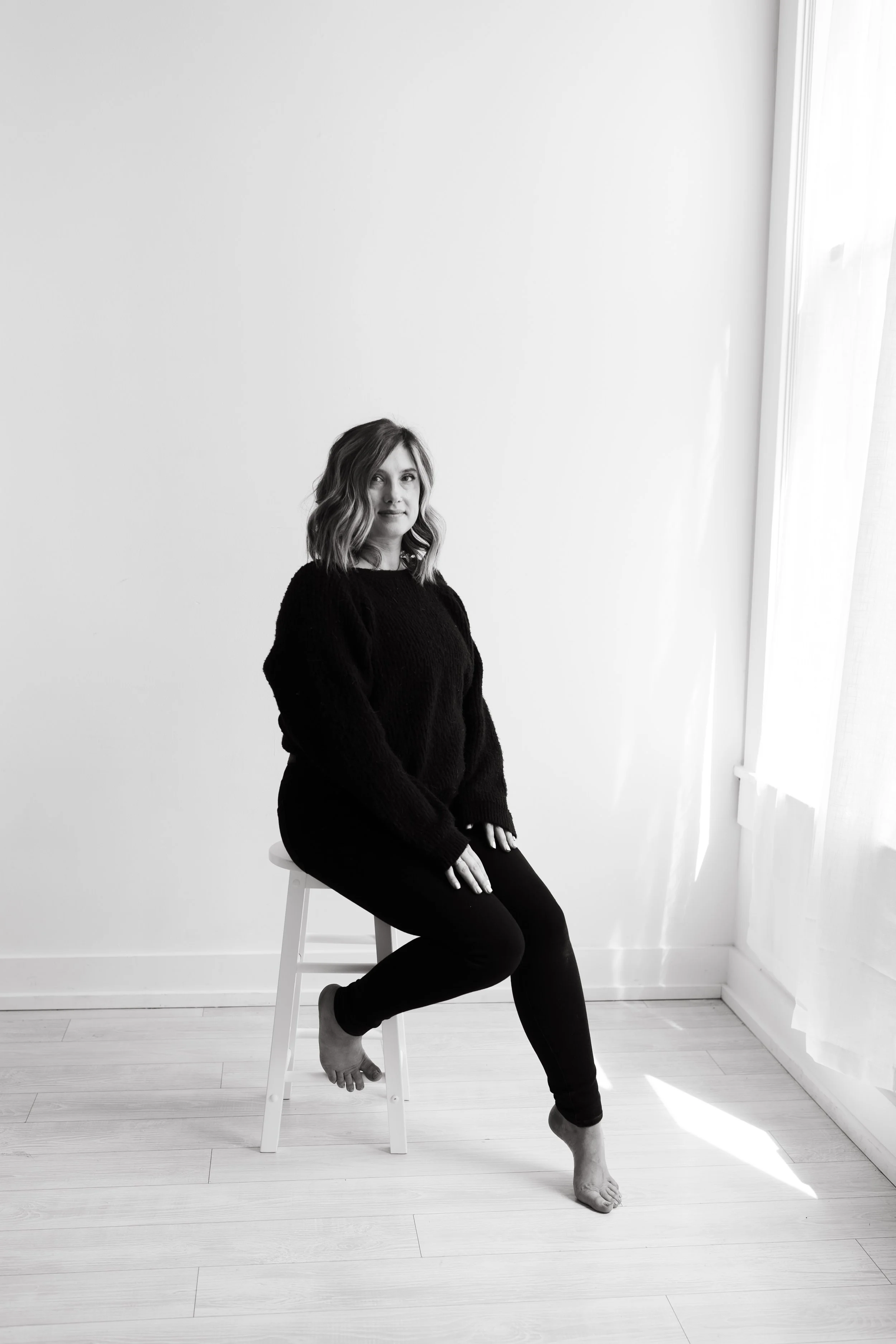 A woman with wavy hair sitting on a white stool in a room with bright natural light, wearing a black sweater and black pants, in black and white.