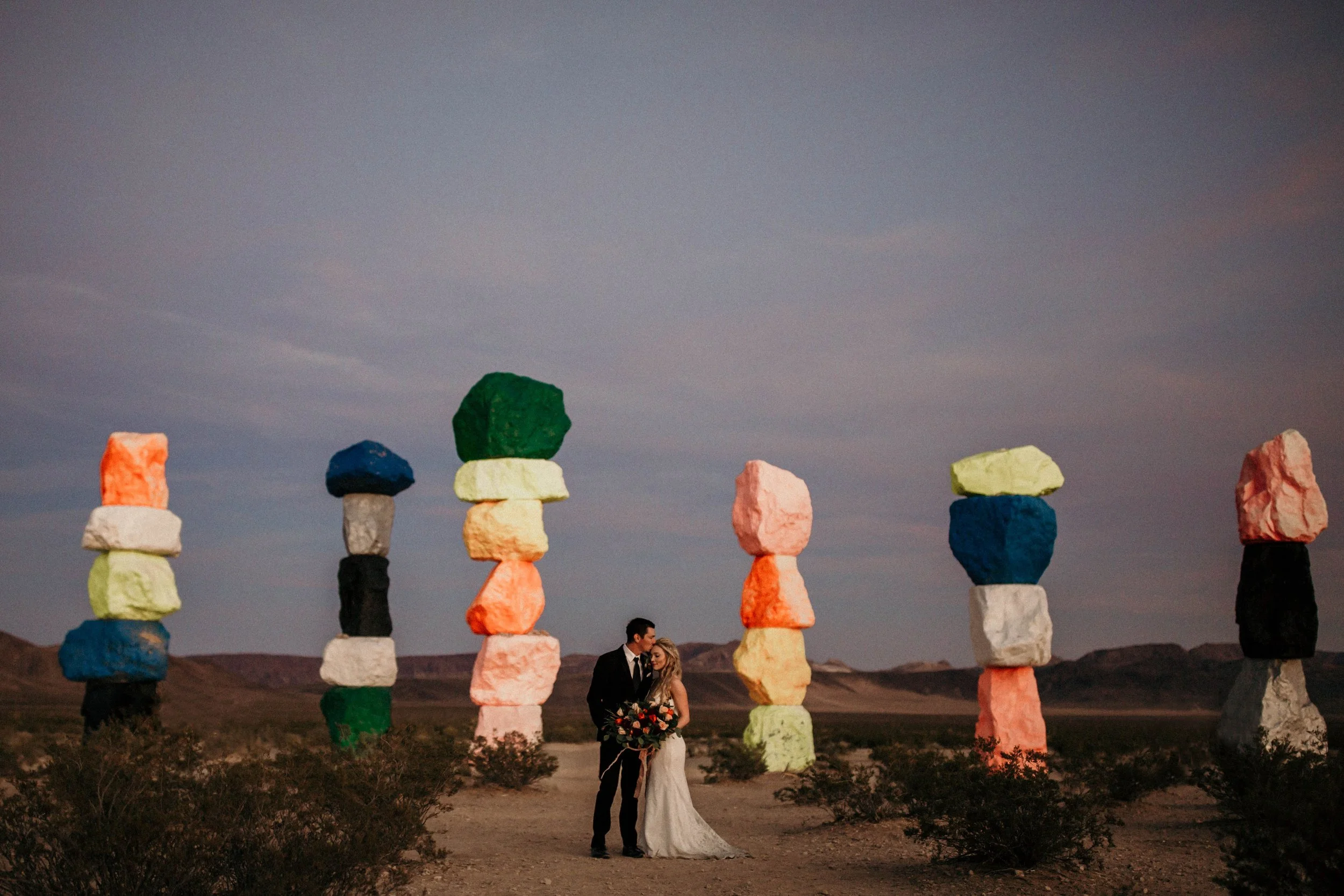 A bride and groom in wedding attire standing close and holding a bouquet, surrounded by colorful stacked rocks in a desert landscape during dusk.