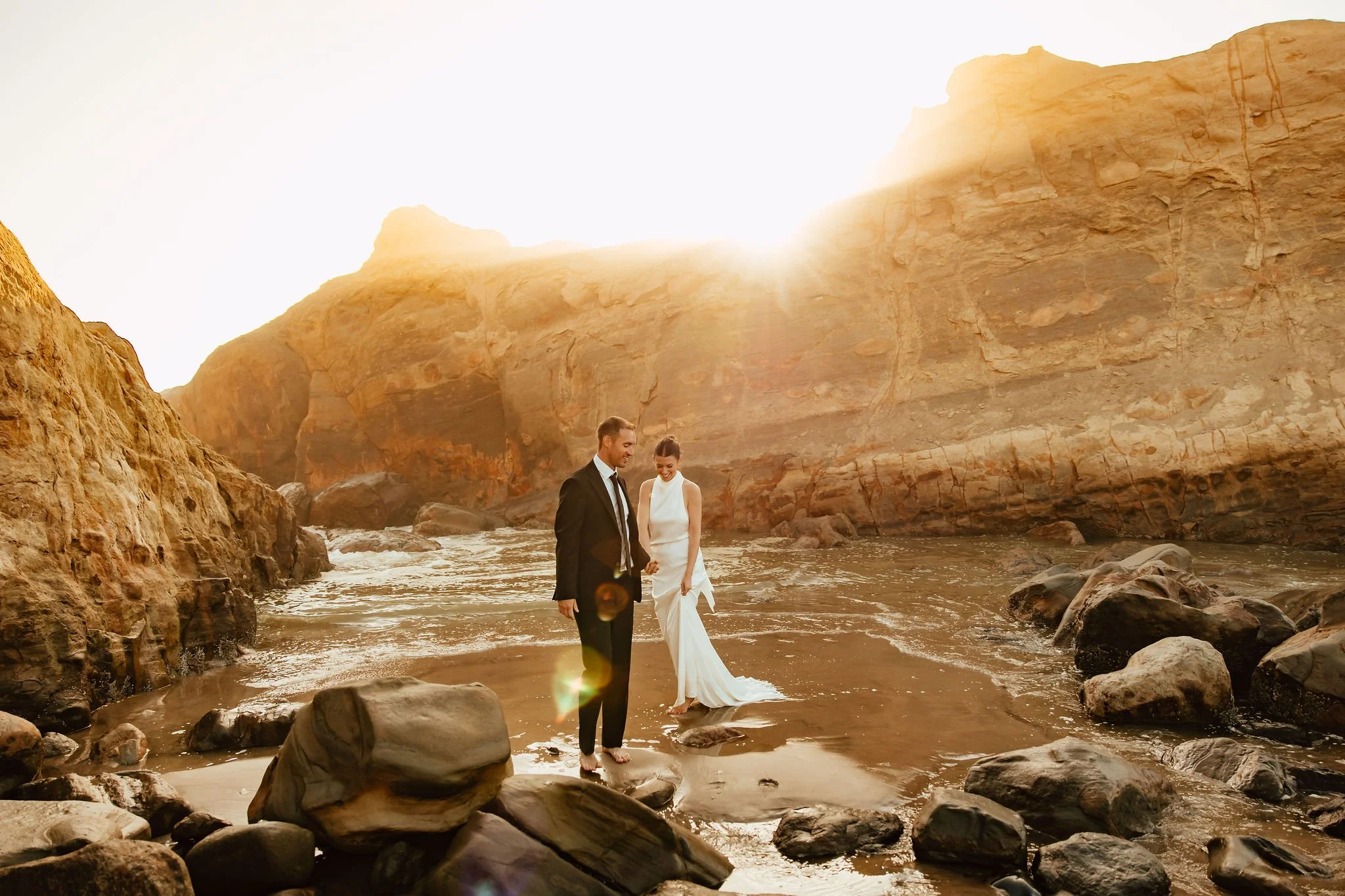A bride and groom in wedding attire walking barefoot on a rocky beach at sunset, holding hands and looking at each other.