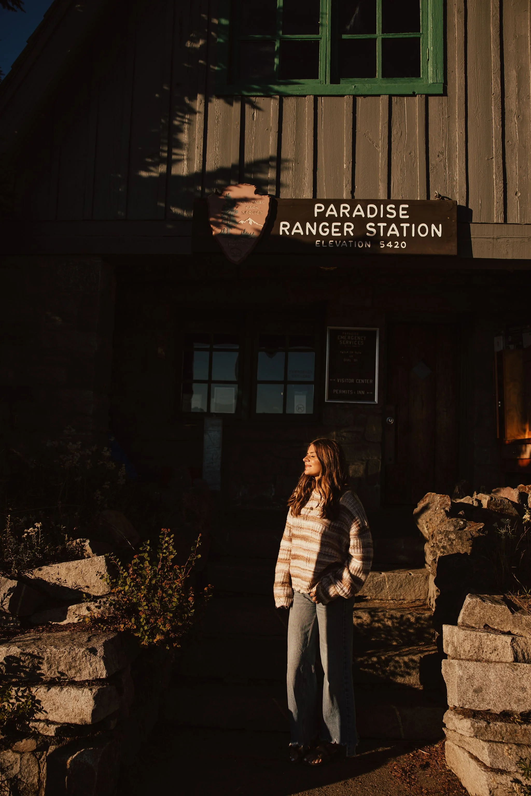 A young woman stands on the steps outside the Paradise Ranger Station, surrounded by rocks and plants, with sunlight illuminating her face.