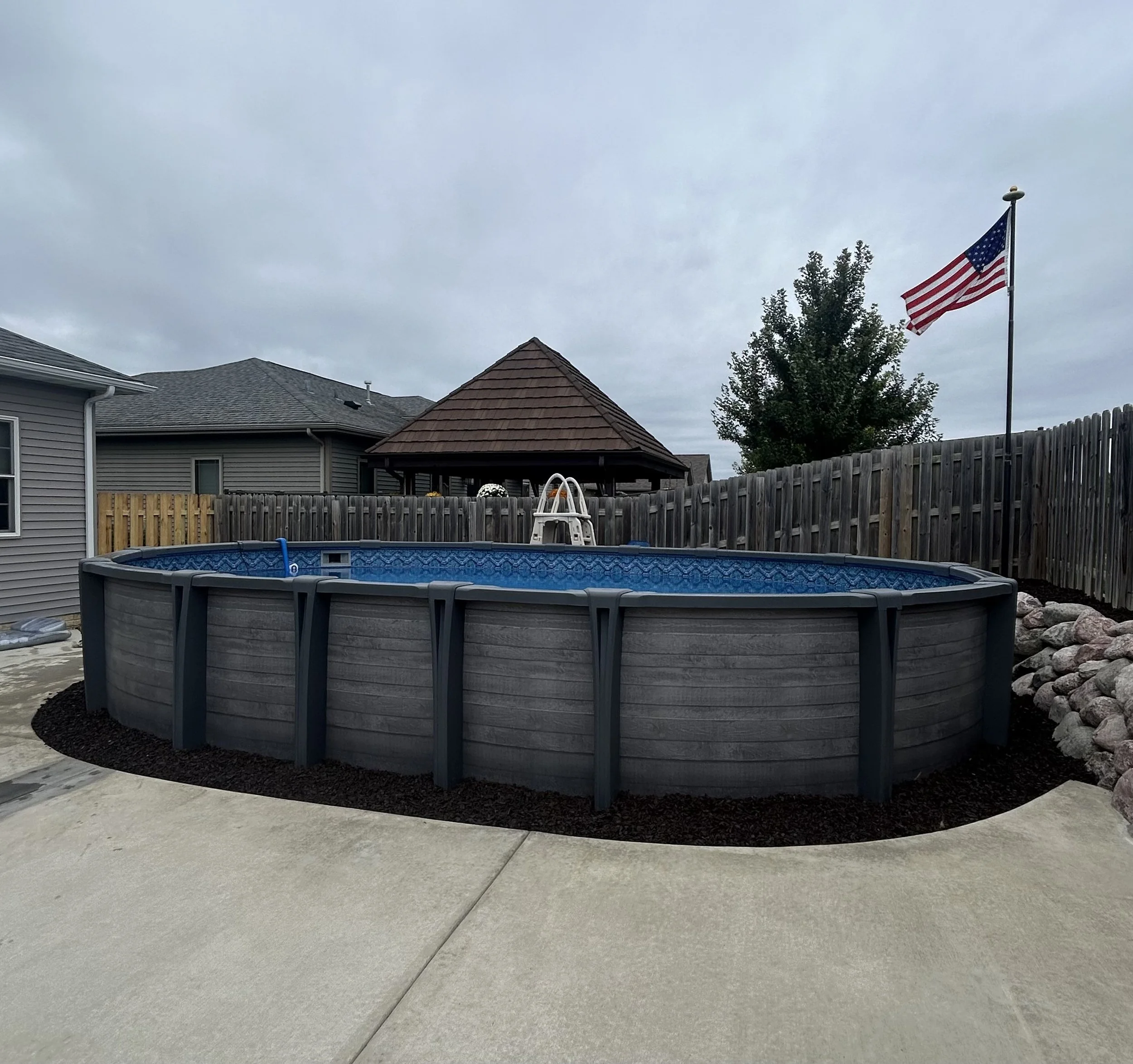 Above-ground swimming pool in a fenced backyard with an American flag and gazebo in the background.