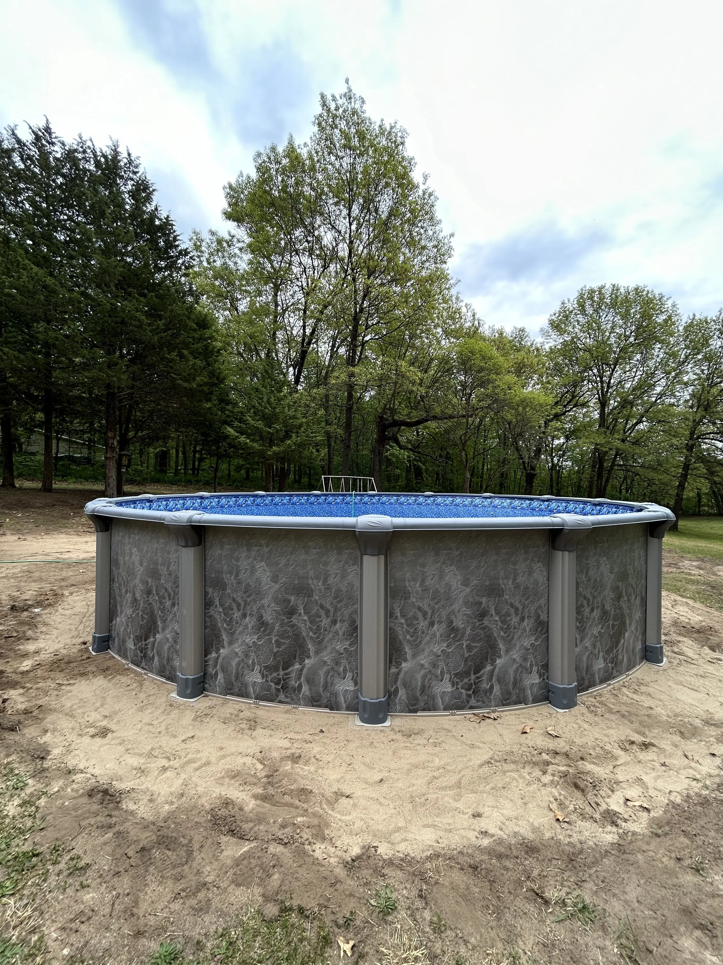 Above-ground pool surrounded by a sandy area and trees in the background.