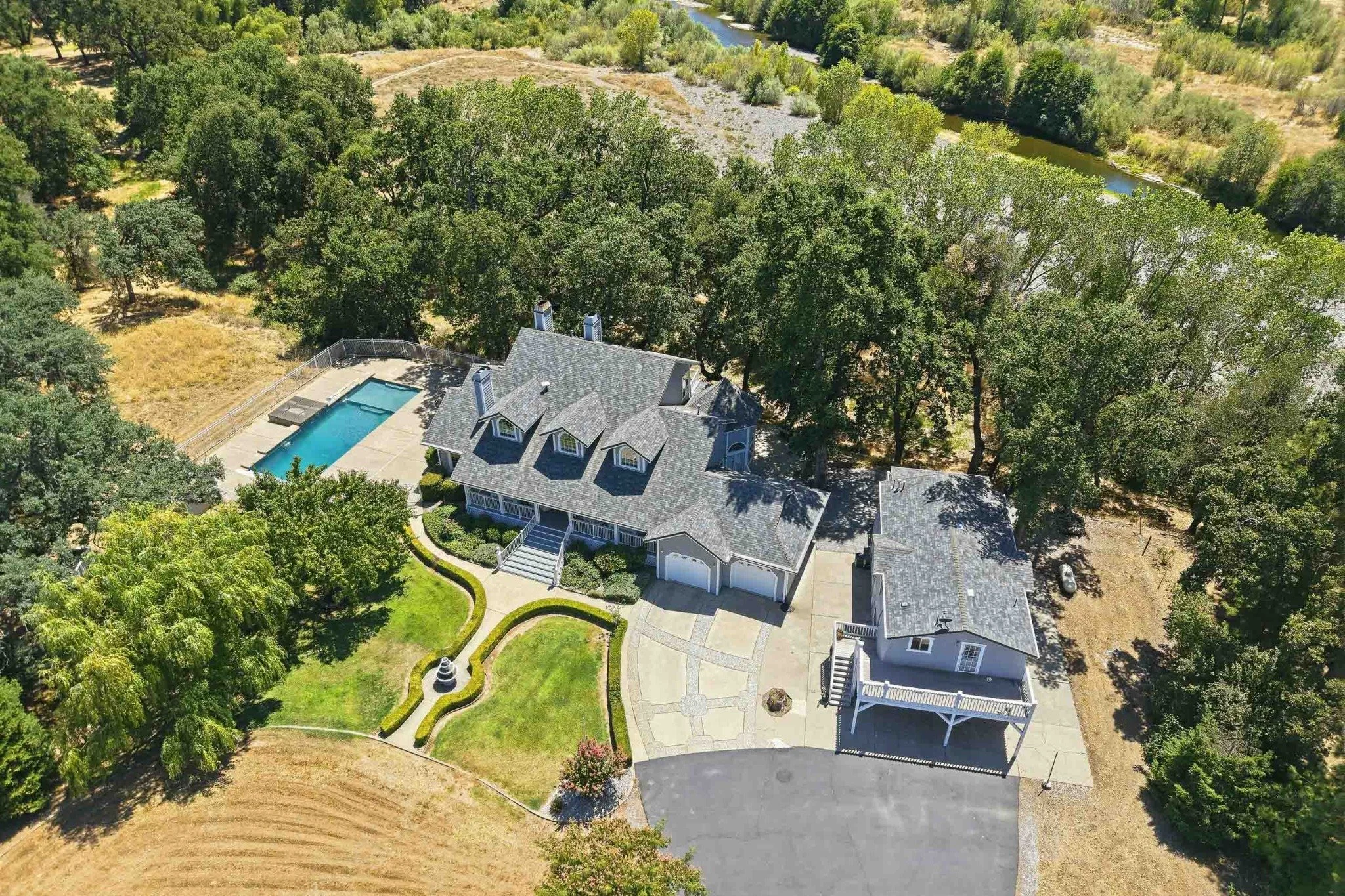 Aerial view of a large estate with a pool, guest house, mature trees, and nearby river.