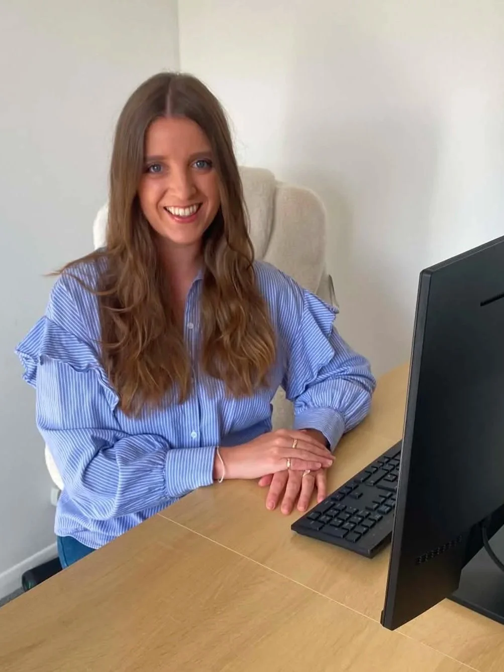 A woman with long, wavy brown hair smiling at the camera, sitting at a desk with a computer monitor and keyboard.