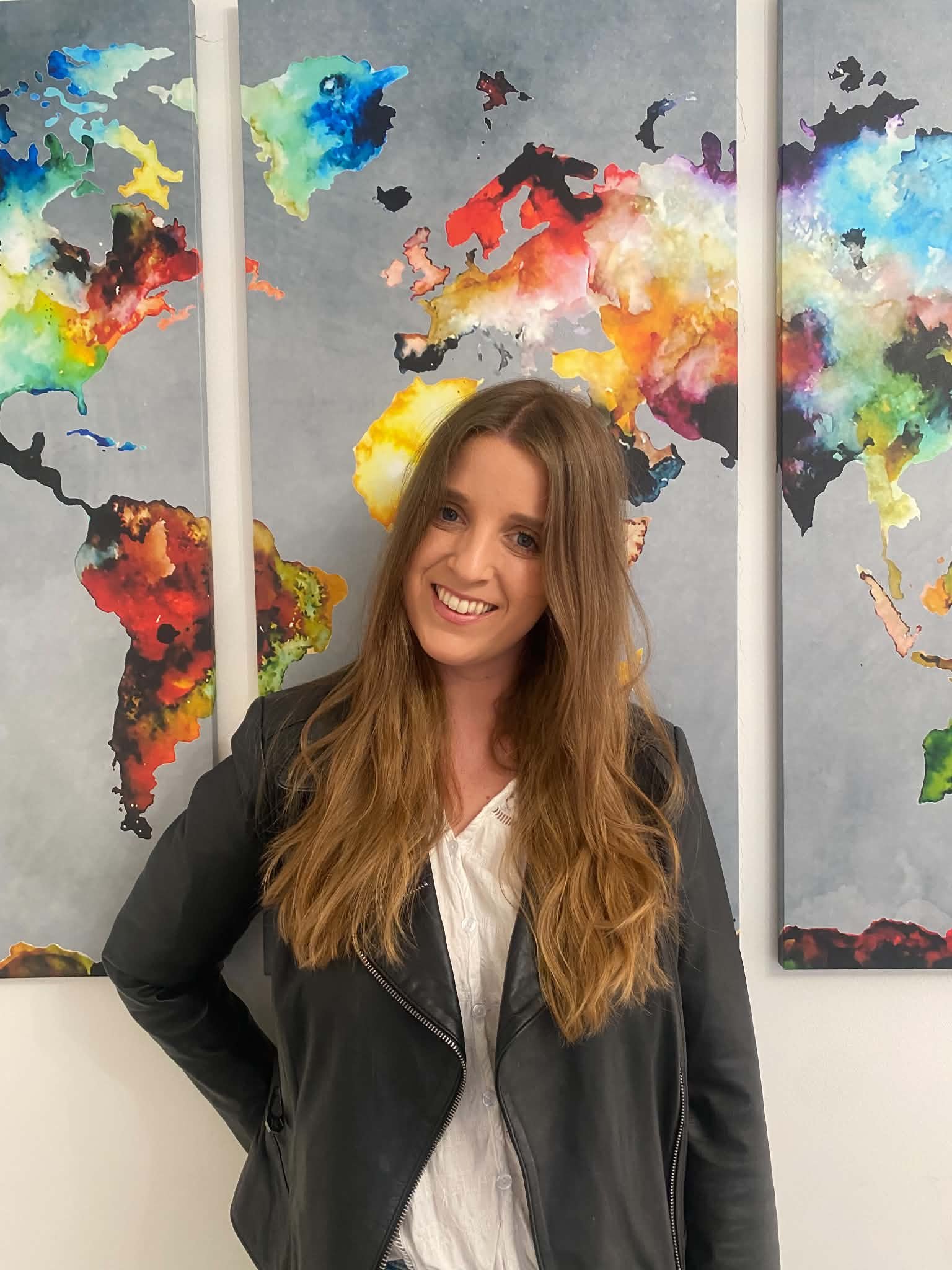 A smiling woman with long reddish-brown hair wearing a black leather jacket standing in front of a colorful, abstract world map mural on a gray background.