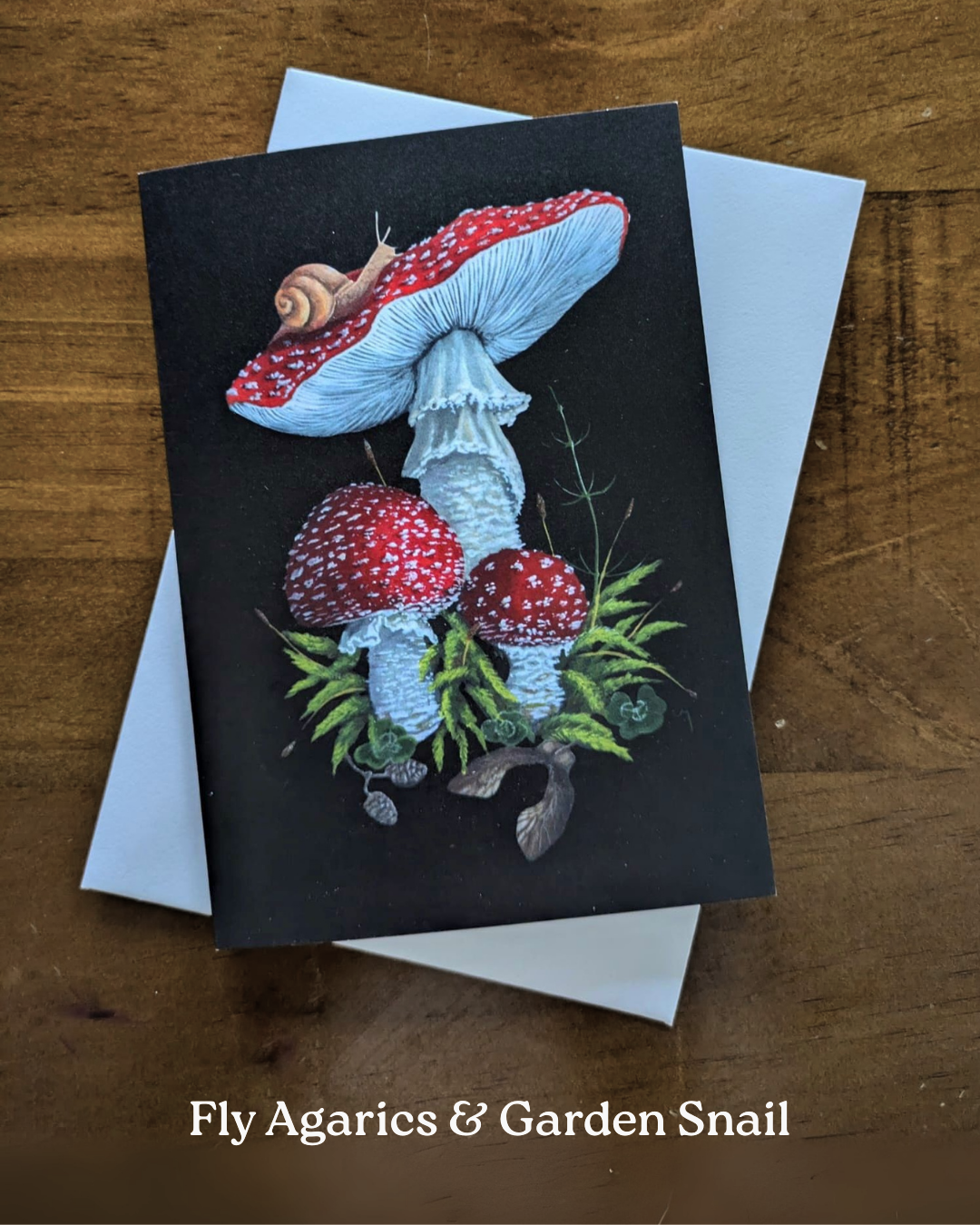 A greeting card with a painted Red Fly Agaric mushrooms & a Garden snail surrounded by moss on a black background.