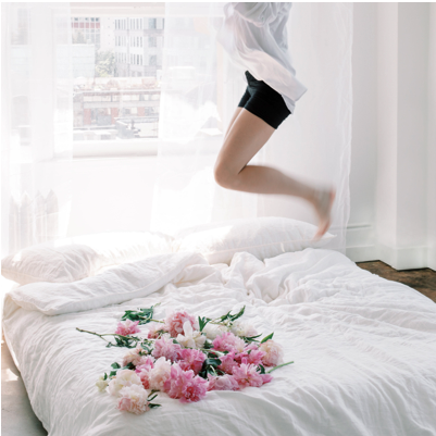 Frau springt voller Energie auf einem hellen Bett mit Pfingstrosen – Darstellung von wiedergewonnener Beckenboden-Kontinenz, Lebensfreude und körperlicher Belastbarkeit.