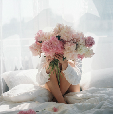 Frau sitzt im hellen Bett und hält einen großen Strauß rosa Pfingstrosen vor ihr Gesicht – Symbol für die sanfte Regeneration und hormonelle Balance im Wochenbett.