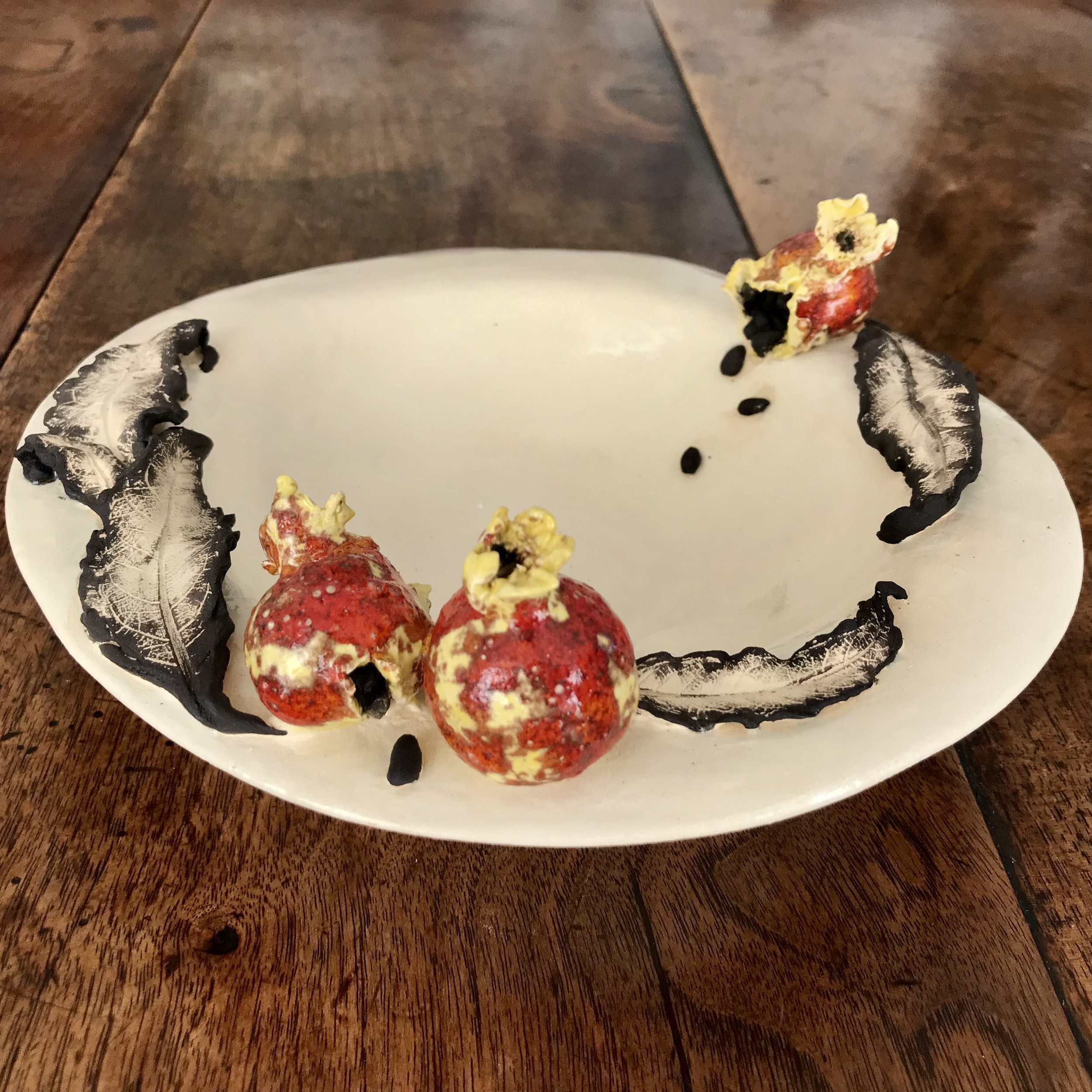 Volcanic Victuals: hand built stoneware bowl with sculpted pomegranates, glazed, beside leaves and seeds brushed with black powder.