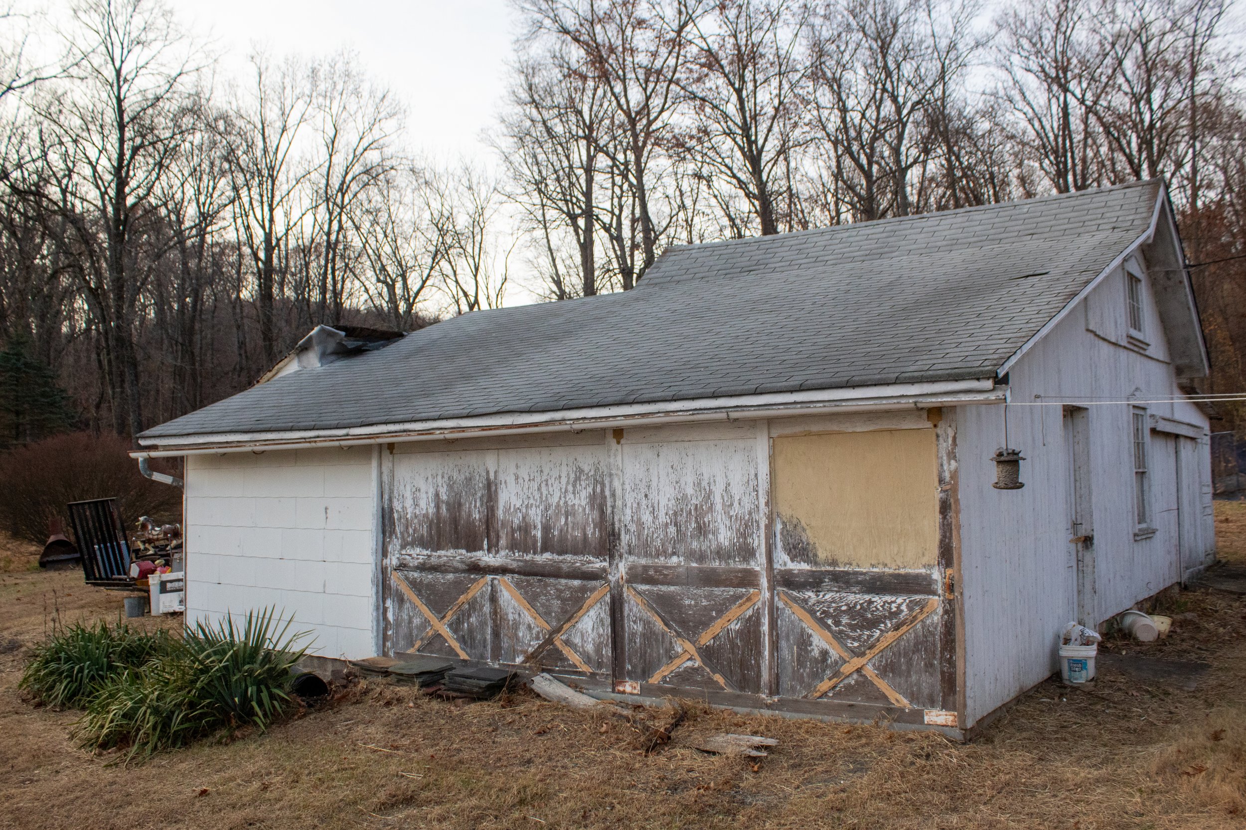 Old wooden shed with weathered paint and a gray shingled roof, surrounded by leafless trees and grass.