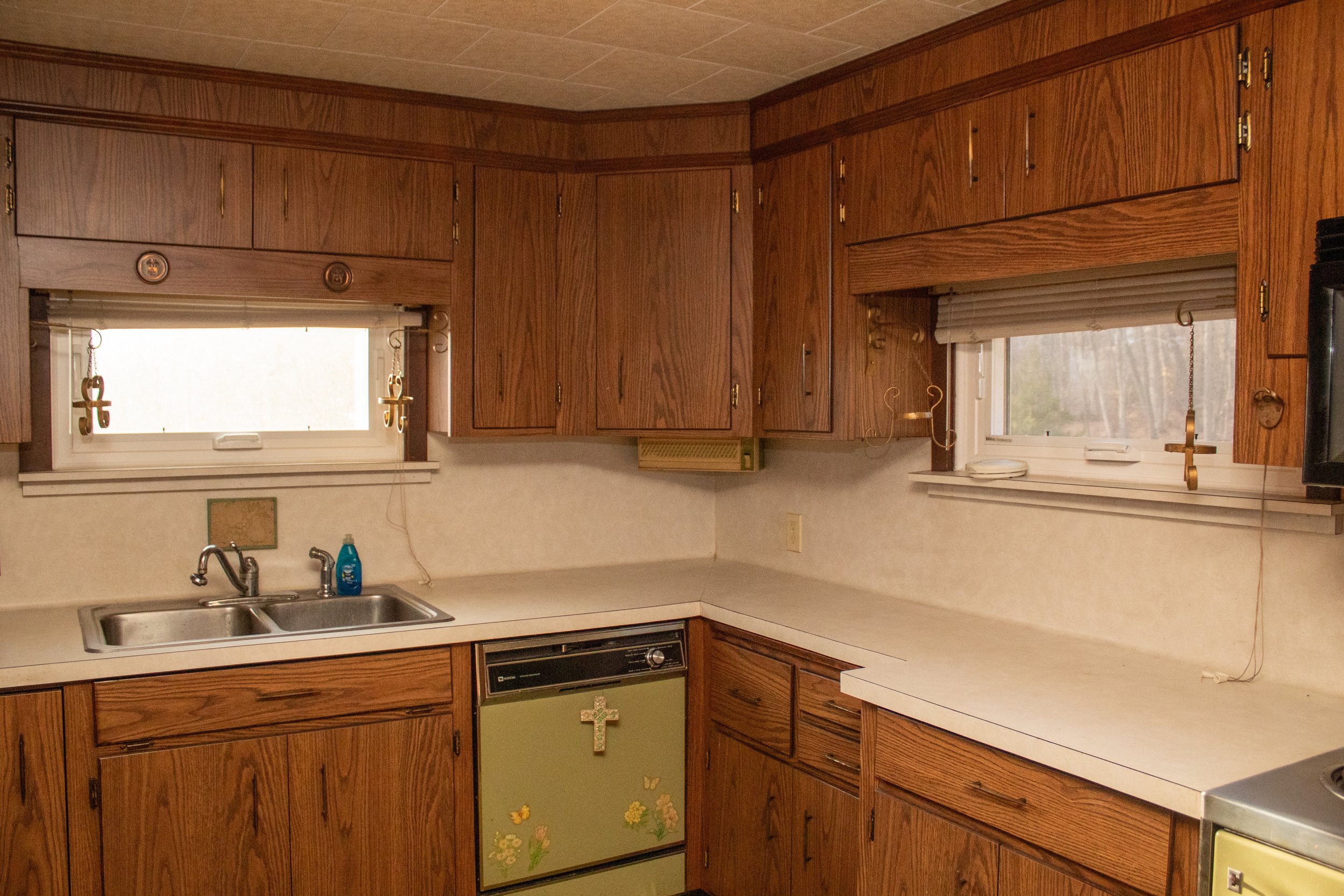 Vintage kitchen with wooden cabinets, white countertops, double sink, a yellow-green dishwasher decorated with floral designs and a cross, multiple windows with blinds, and various hanging items.