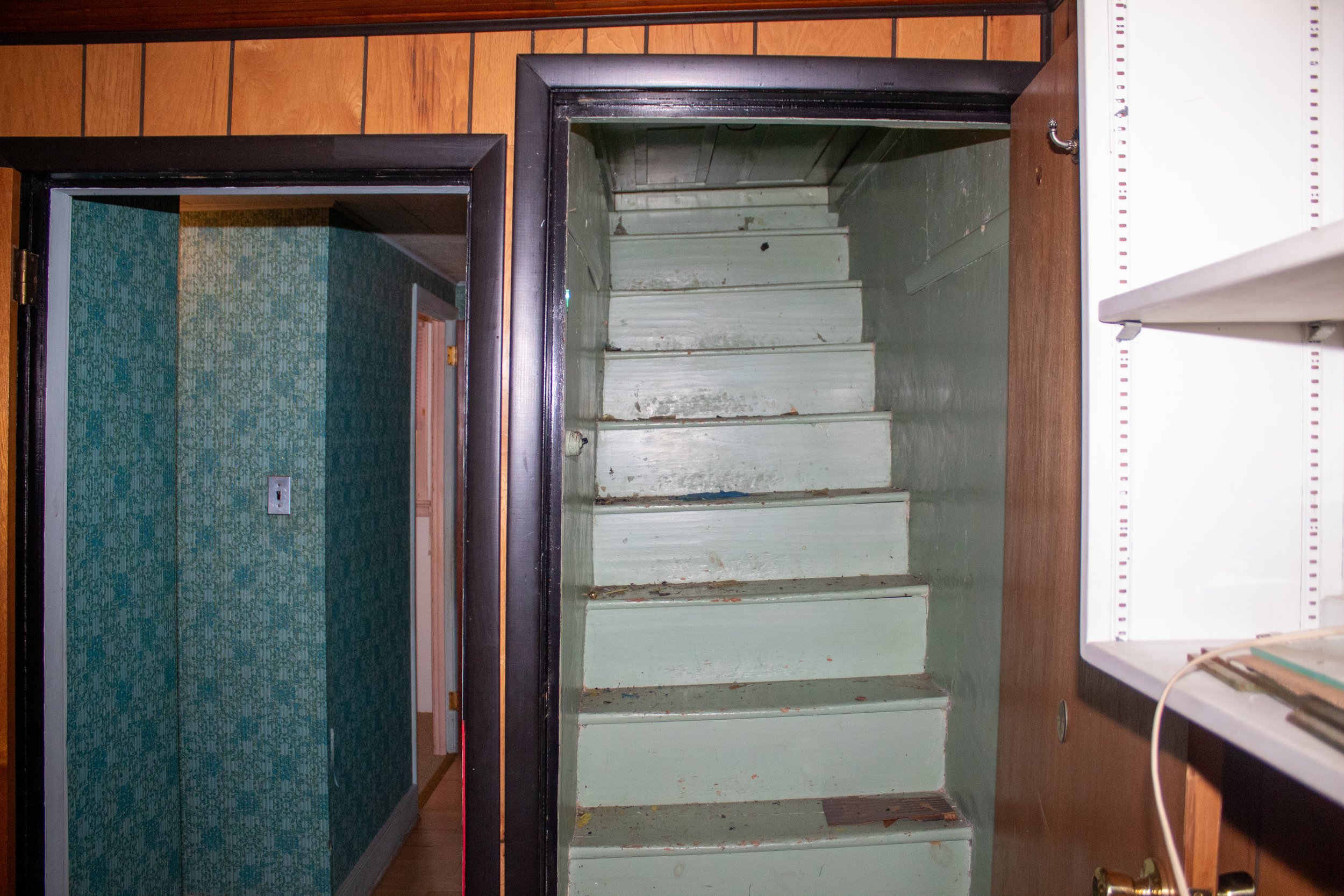 Old narrow staircase in a wooden paneled room with adjacent wallpapered hallway.