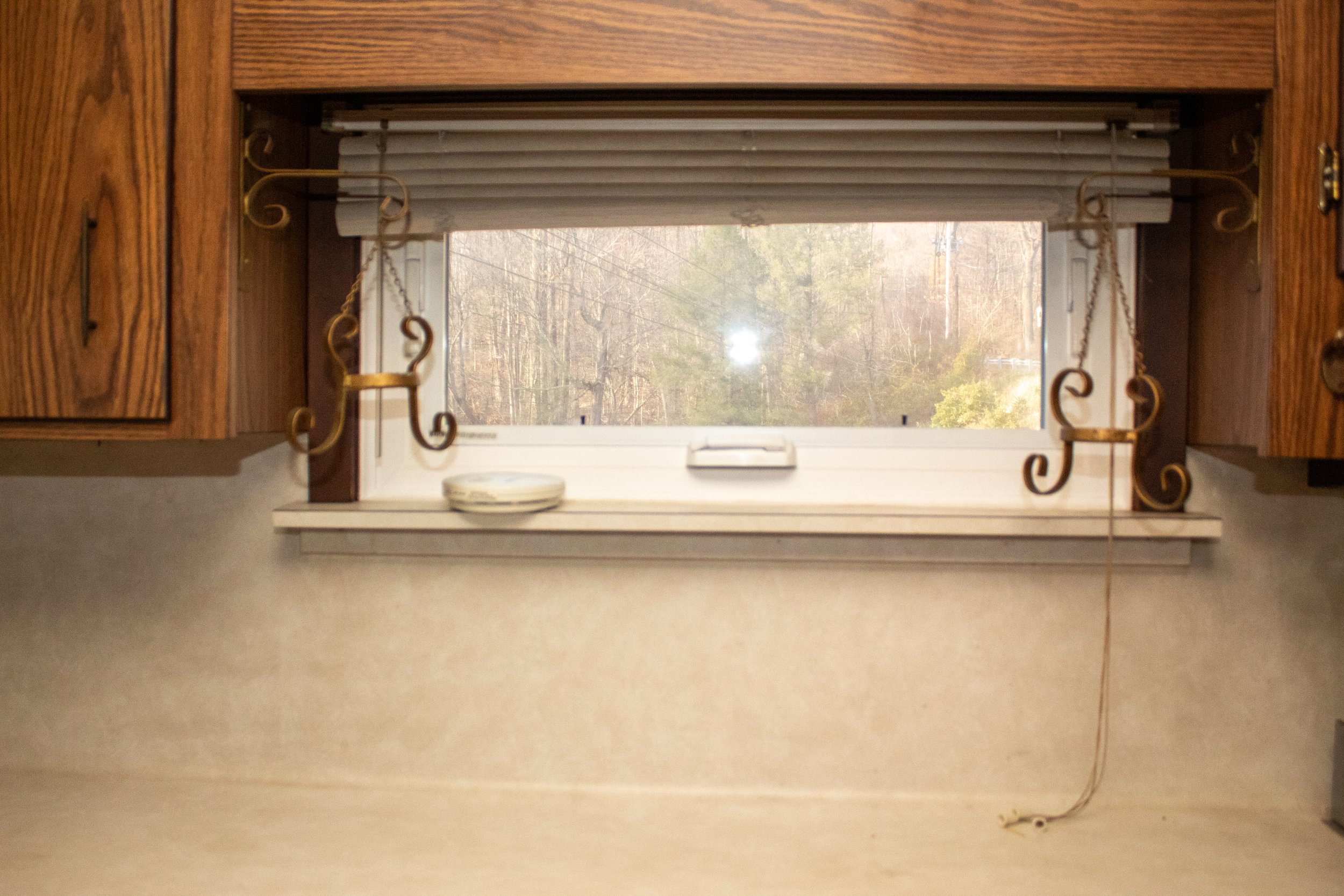 Kitchen counter with a window above, featuring wooden cabinets on both sides. The window has horizontal blinds partially open, showing trees outside. A small dish and decorative metal holders are on the windowsill.
