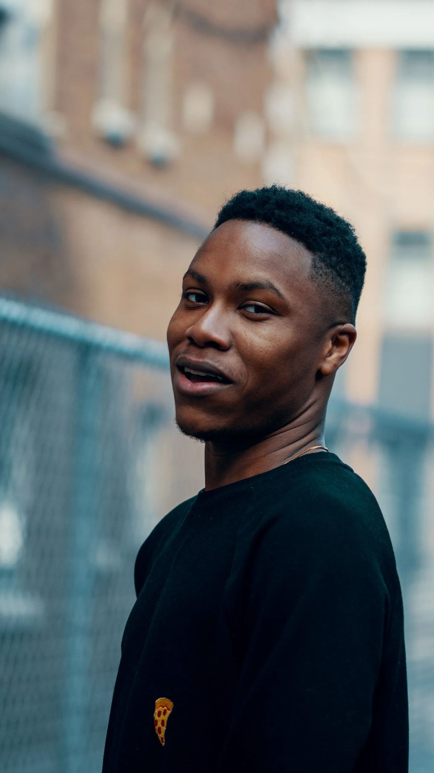 Person smiling outdoors near chain-link fence and brick building, wearing a black shirt with a small pizza slice graphic.