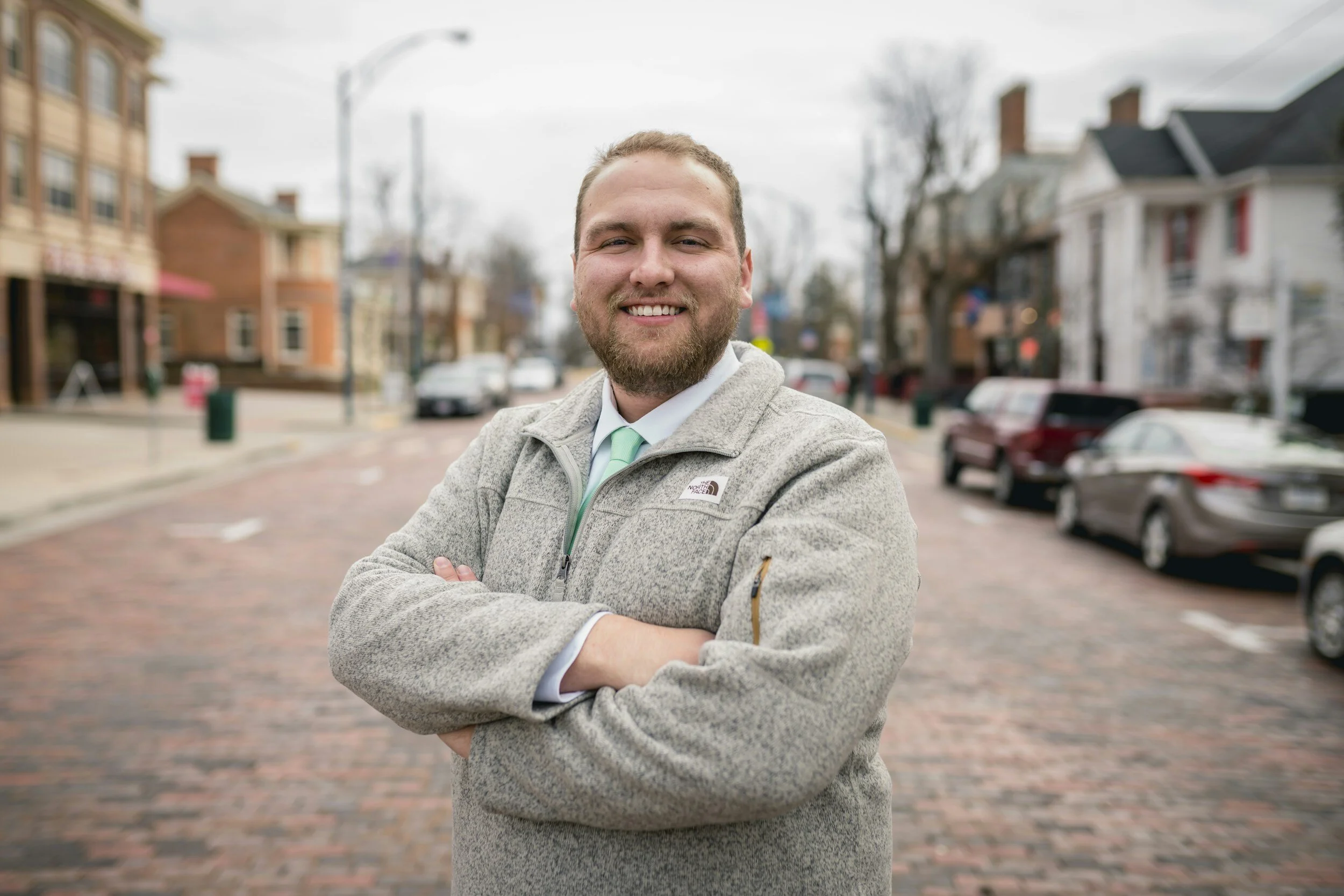 Man smiling with arms crossed on a brick street, wearing a gray jacket and tie, with blurred background of buildings and parked cars.