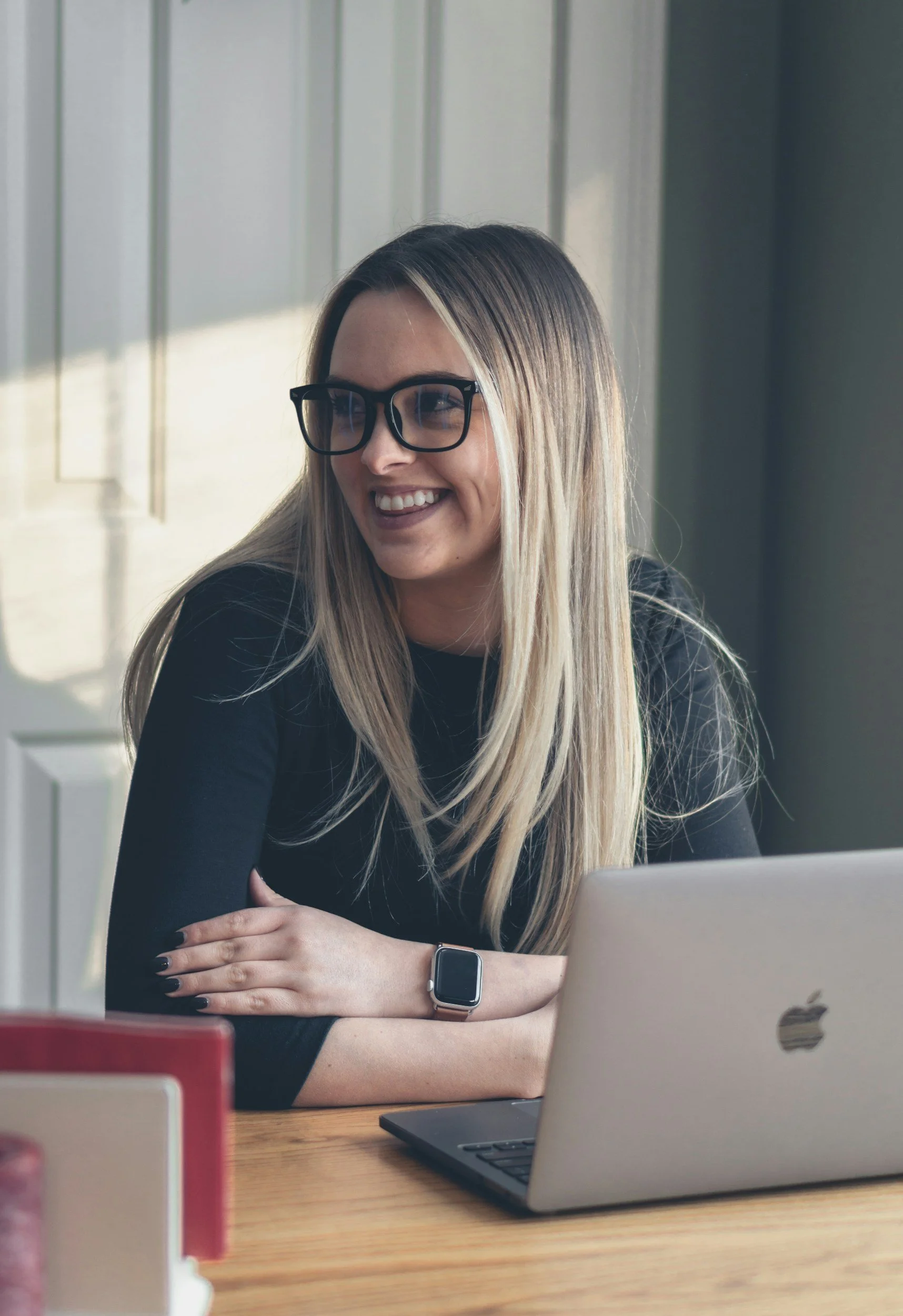 Woman wearing glasses smiling while sitting at a desk with a laptop and a smartwatch.