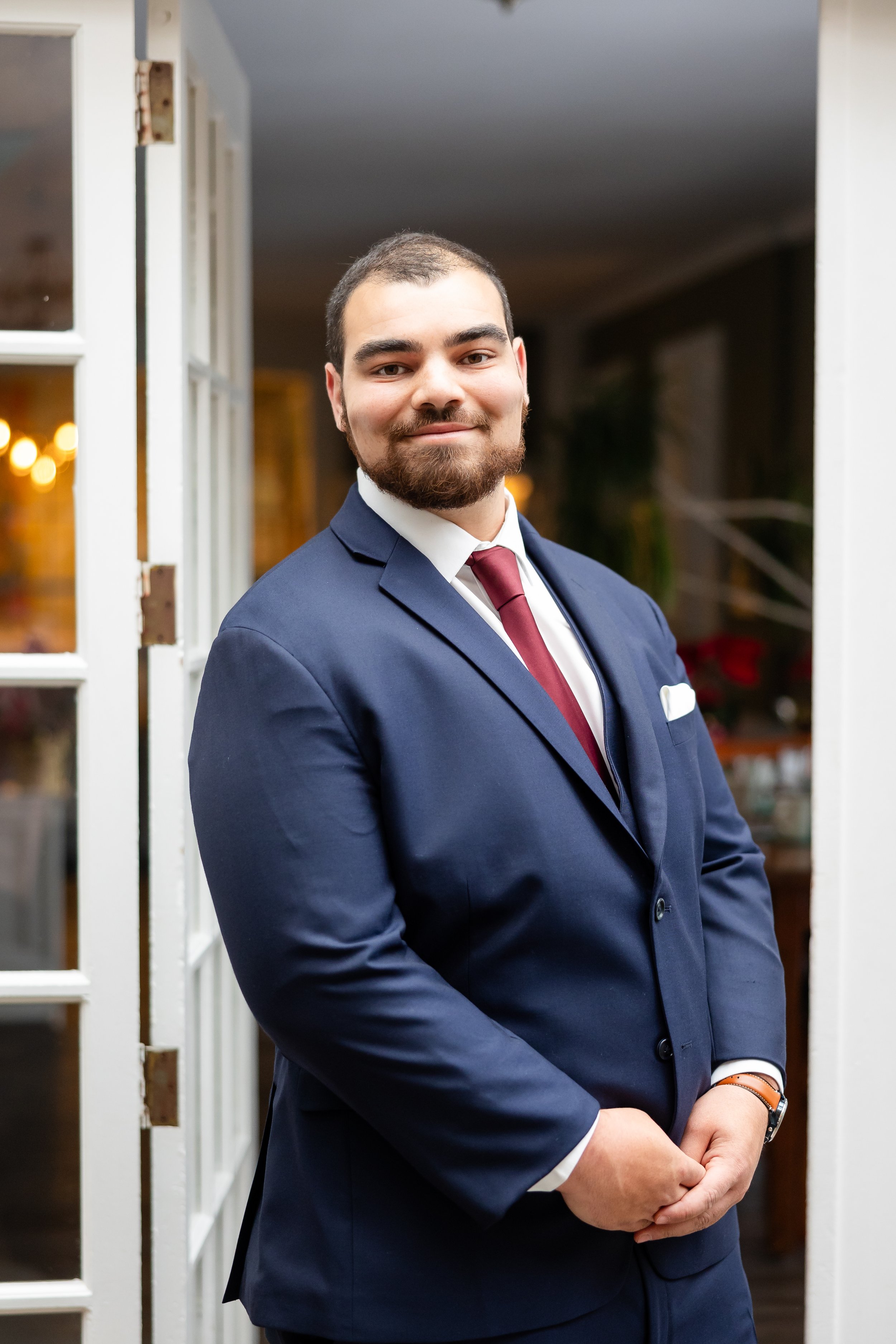 A man in a navy suit with a burgundy tie and boutonniere stands indoors against a wall made of multicolored wooden planks, smiling slightly toward the camera, with soft natural light coming from a nearby window.