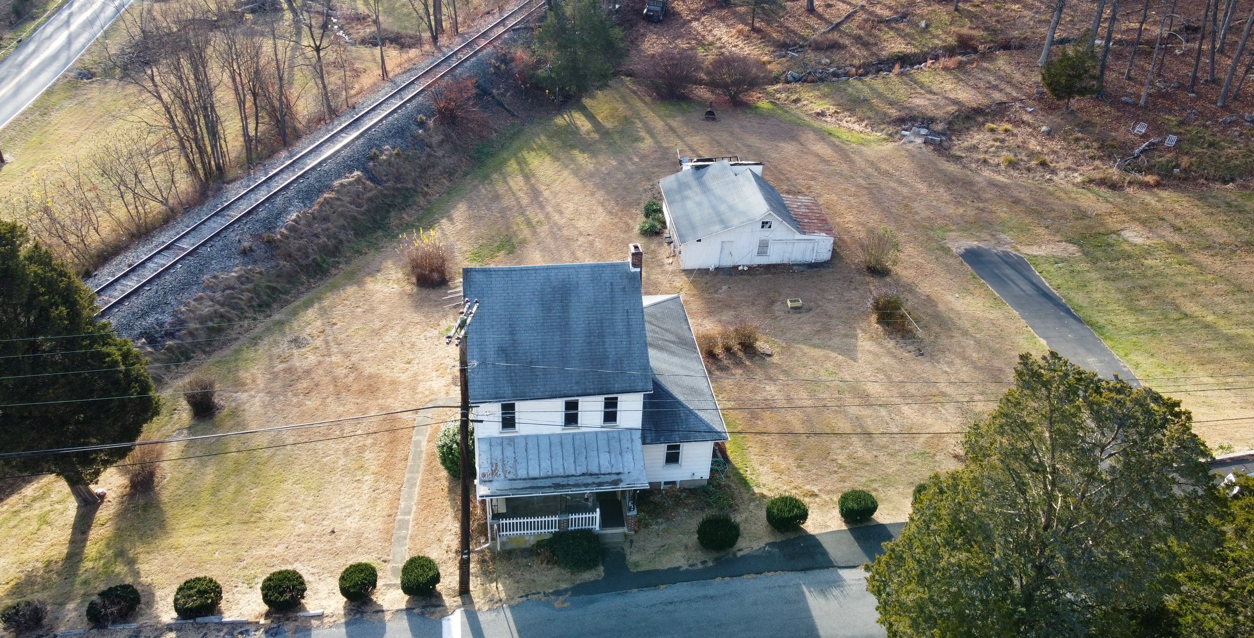 Aerial view of a rural landscape featuring an old white house with a roof and porch, a small detached garage, grassy yard, and a railroad track running alongside it. Trees and a driveway are also visible.