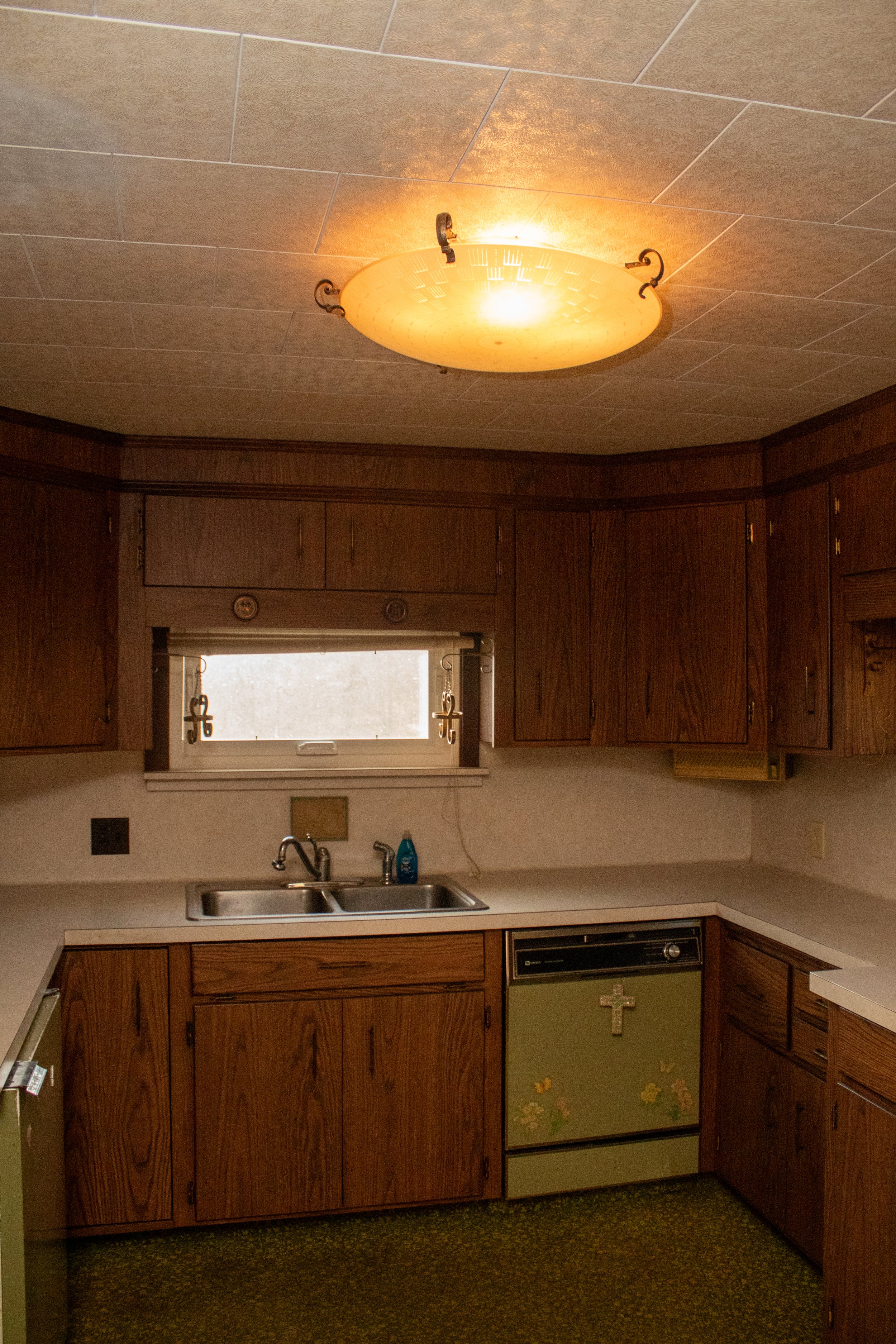 Vintage kitchen with wooden cabinets, double sink, green dishwasher, and overhead light fixture.