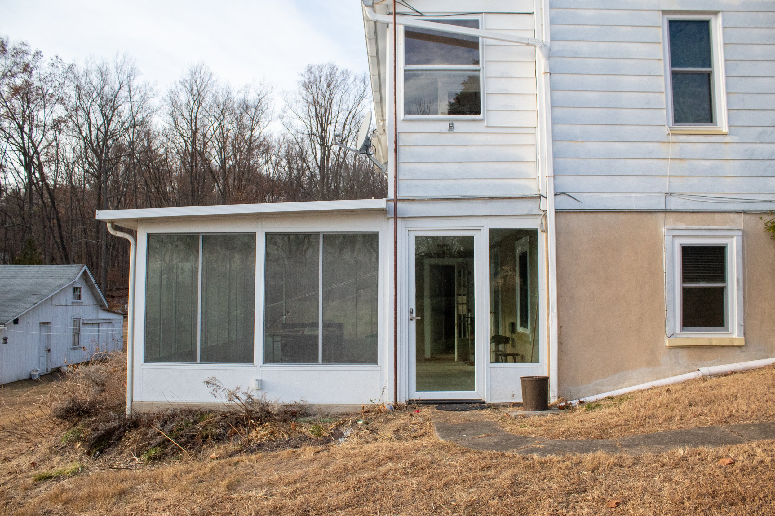 Exterior view of an enclosed porch attached to a house, with sliding glass doors and surrounding windows, situated on a dry, grassy area with bare trees in the background.