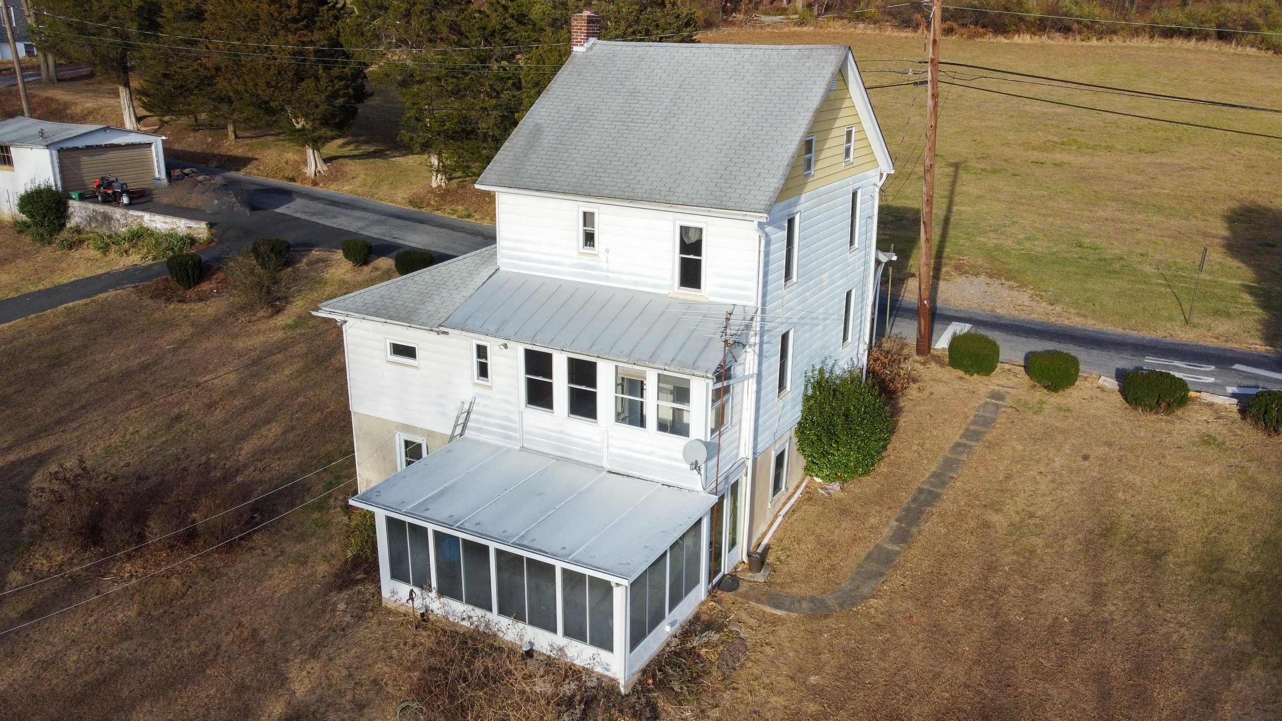 Aerial view of a white two-story house with a gray roof, surrounded by grass, trees, and a driveway. There is a small garage with a vehicle nearby.