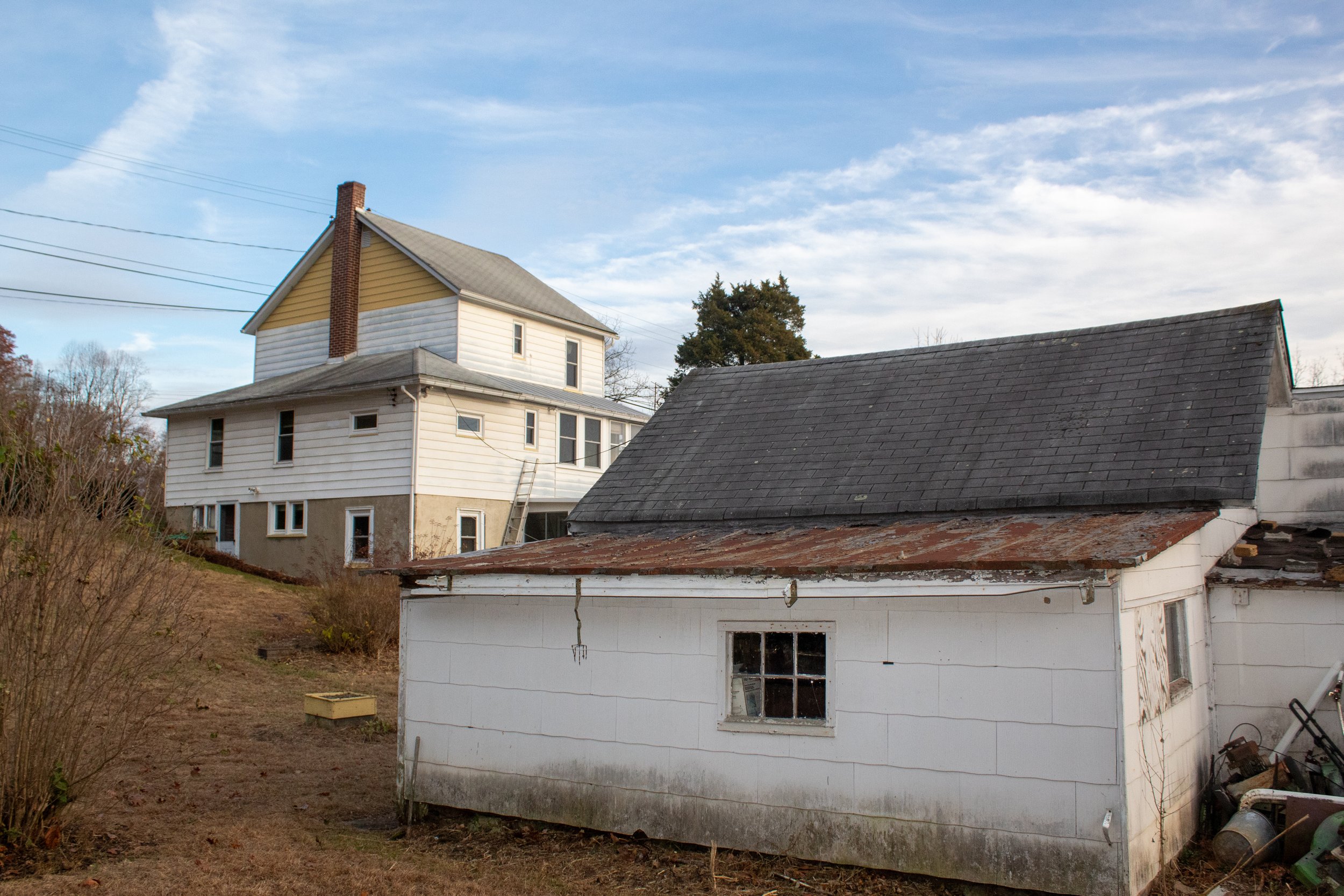 Two-story house with a gabled roof and a smaller, weathered white shed in the foreground, set in a rural landscape.
