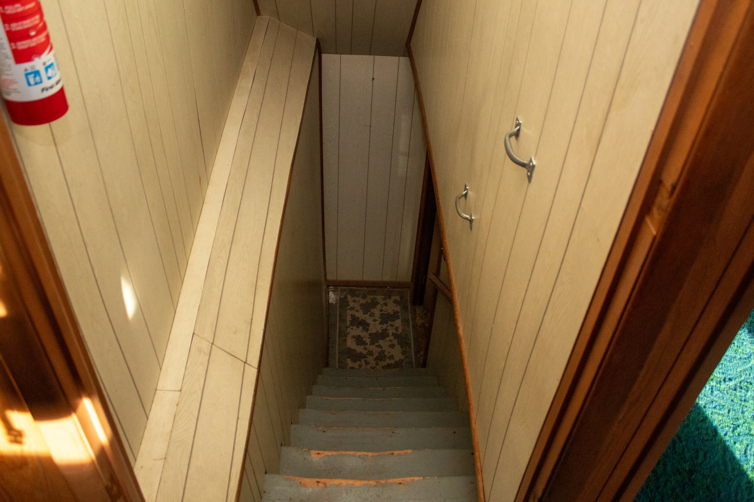 Narrow wooden staircase with handrails leading to a lower level, surrounded by wood-paneled walls, and a fire extinguisher attached to the wall.