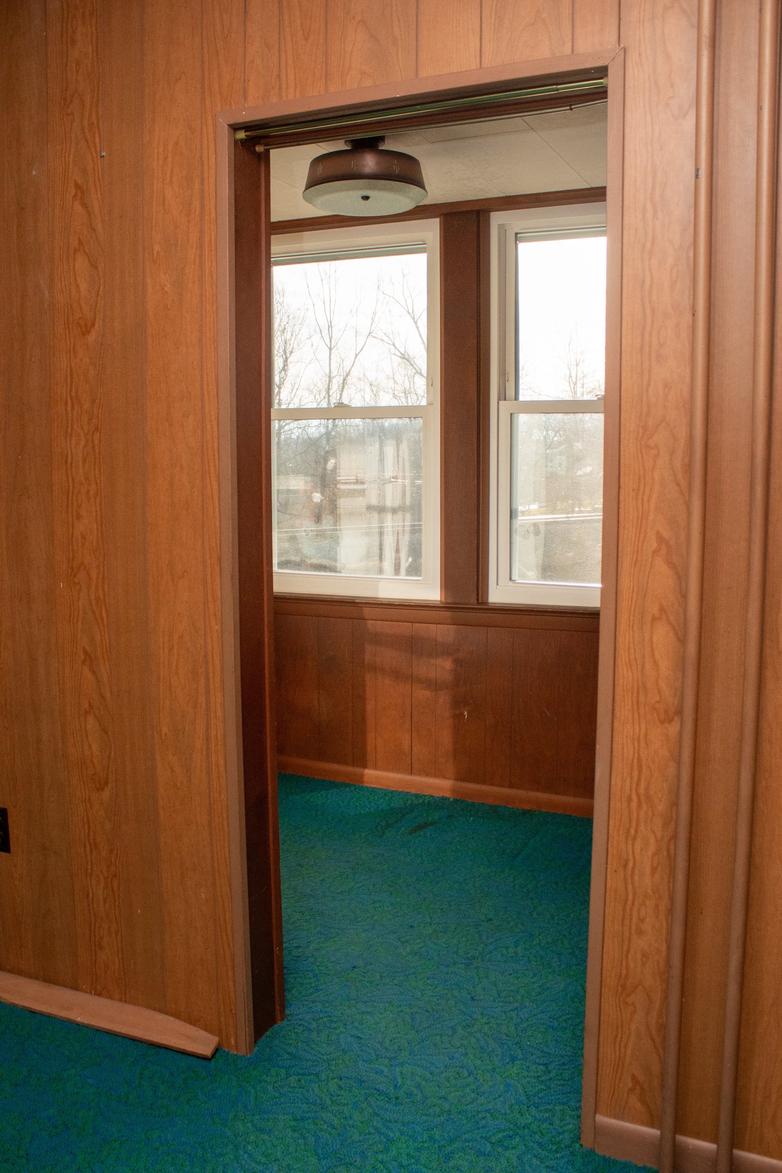 Interior room with wooden paneling, doorway, blue-green carpet, and double windows showing an outdoor view.