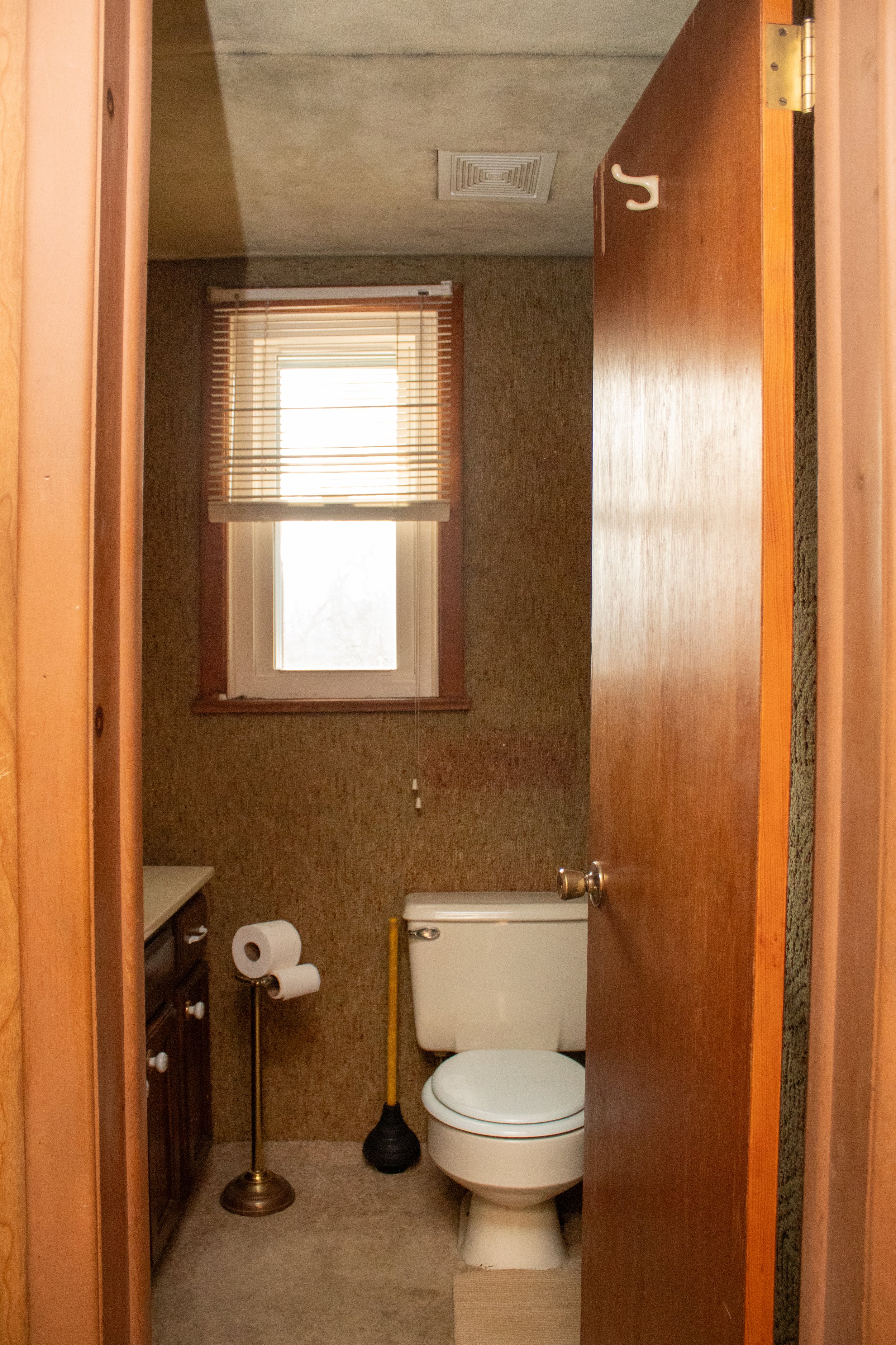 Small bathroom with toilet, toilet paper holder, and plunger, with window blinds above.