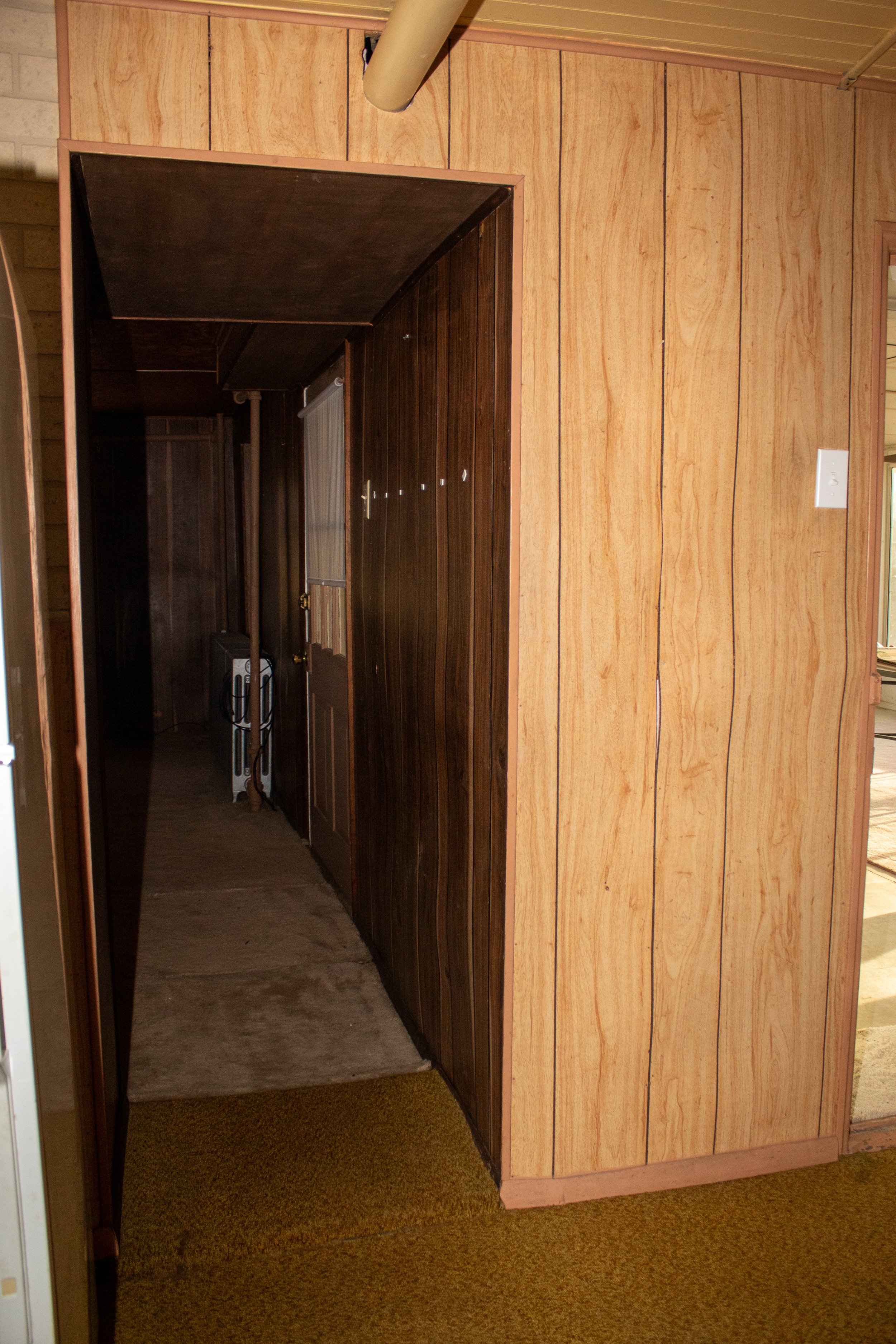 Wood-paneled hallway with a closed door and a narrow passage, featuring contrasting light and dark wood panels and a yellow-brown carpet.
